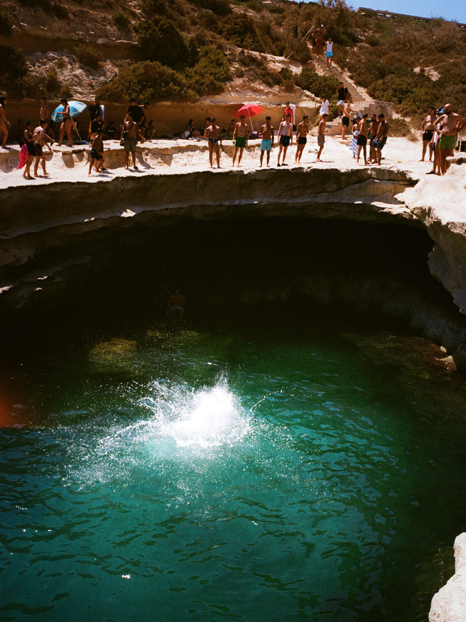 People gather around a natural freshwater pool with a diving board, on a rocky cliffside, during a sunny day.