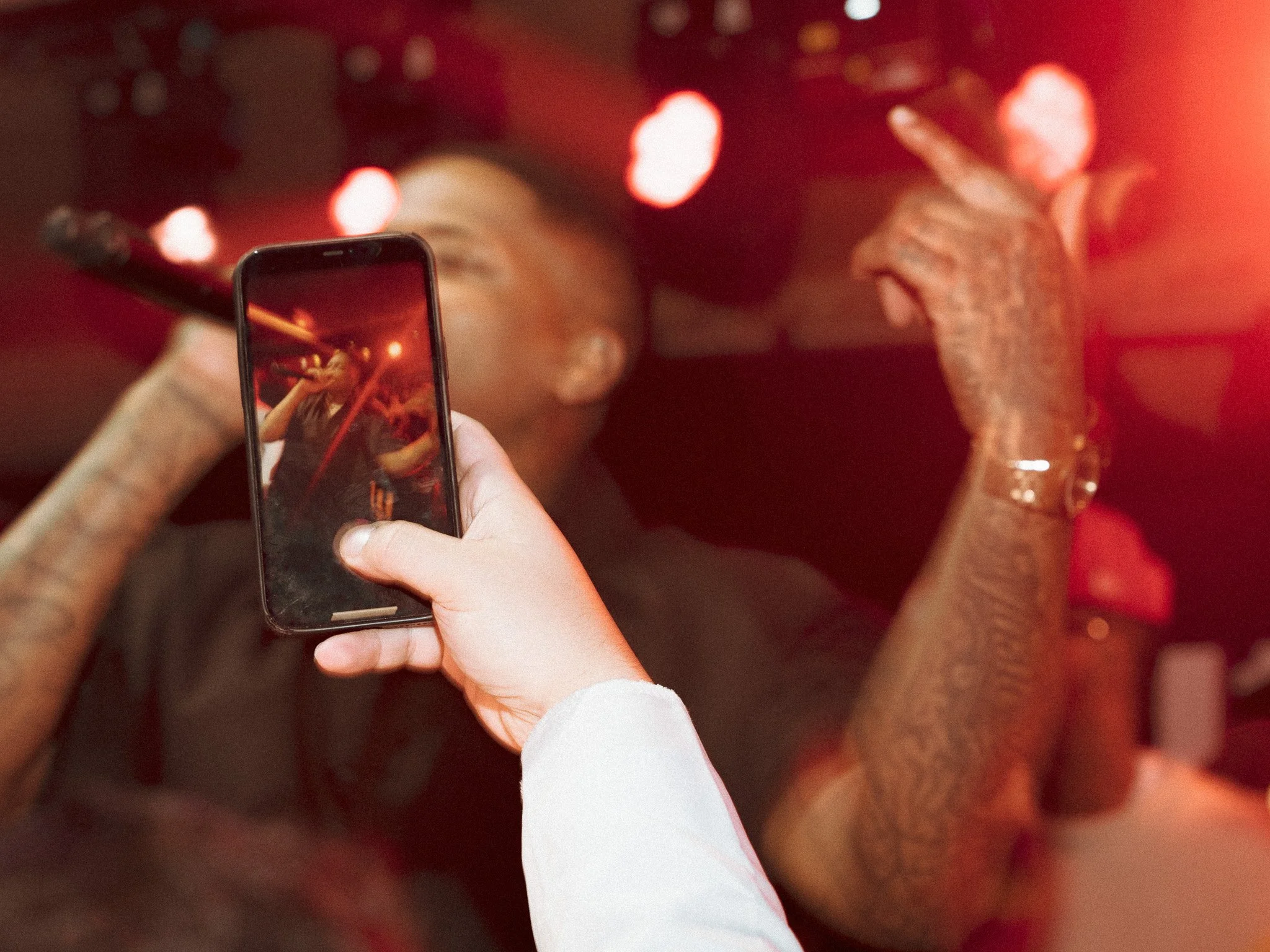 A person with tattoos is on stage holding a microphone and performing, while another person is taking a photo or video using a smartphone in a red-lit environment.