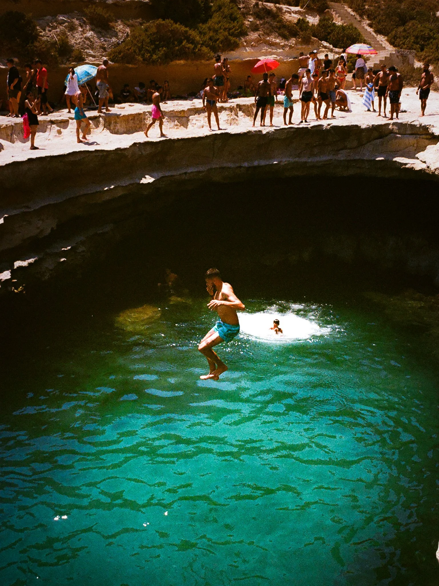 People enjoying swimming and jumping into a natural pool under a rock formation, with onlookers using umbrellas and walking along the edge.