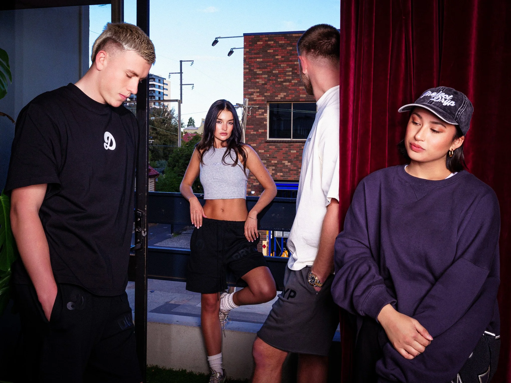 Four young adults inside a room, with one woman standing confidently in front of a window outside and three people near a red curtain, one in a cap and streetwear, another in a white shirt, and another in a black shirt.