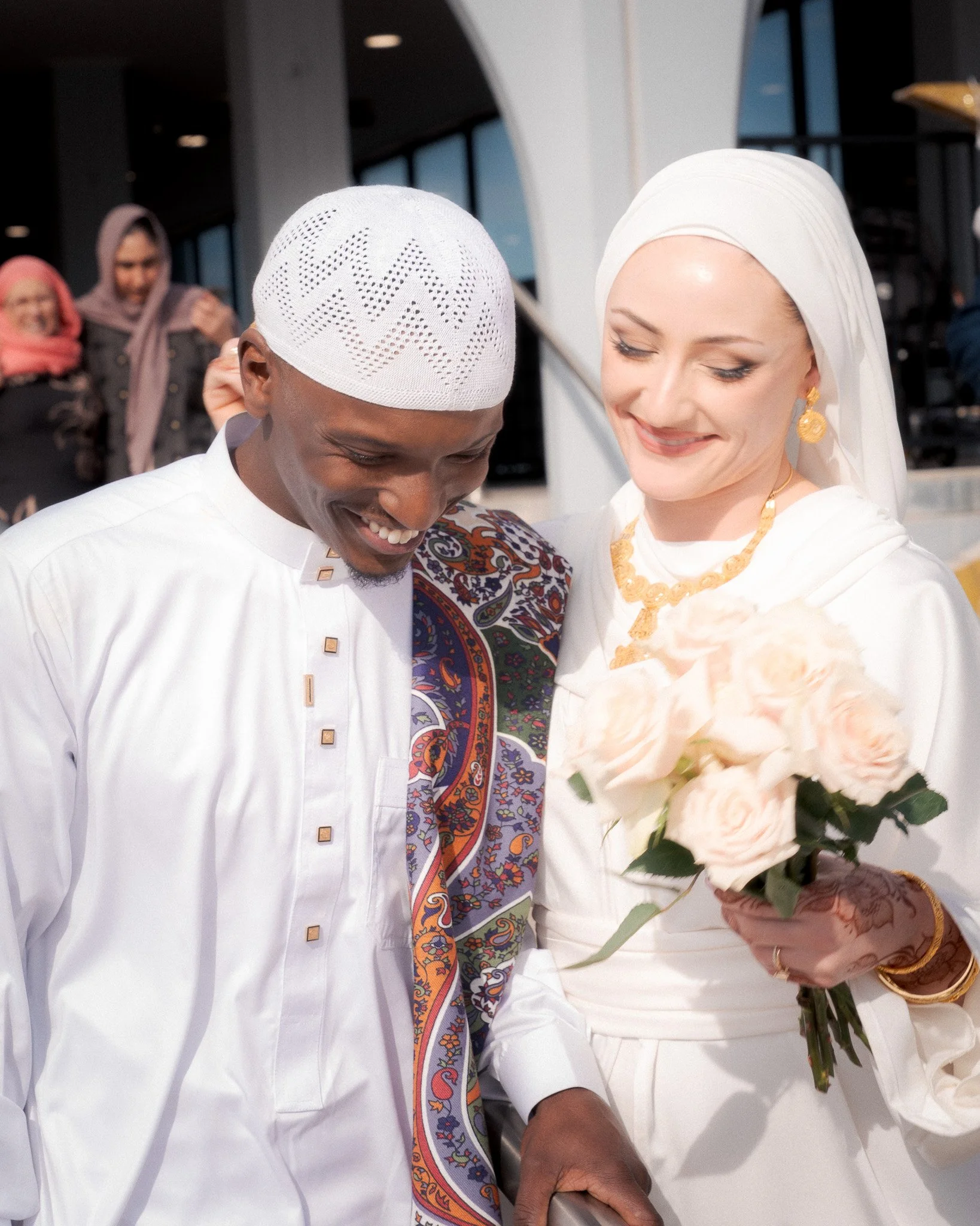 A smiling couple dressed in traditional white attire, with the woman holding a bouquet of light pink roses, standing near large windows with a few onlookers in the background.