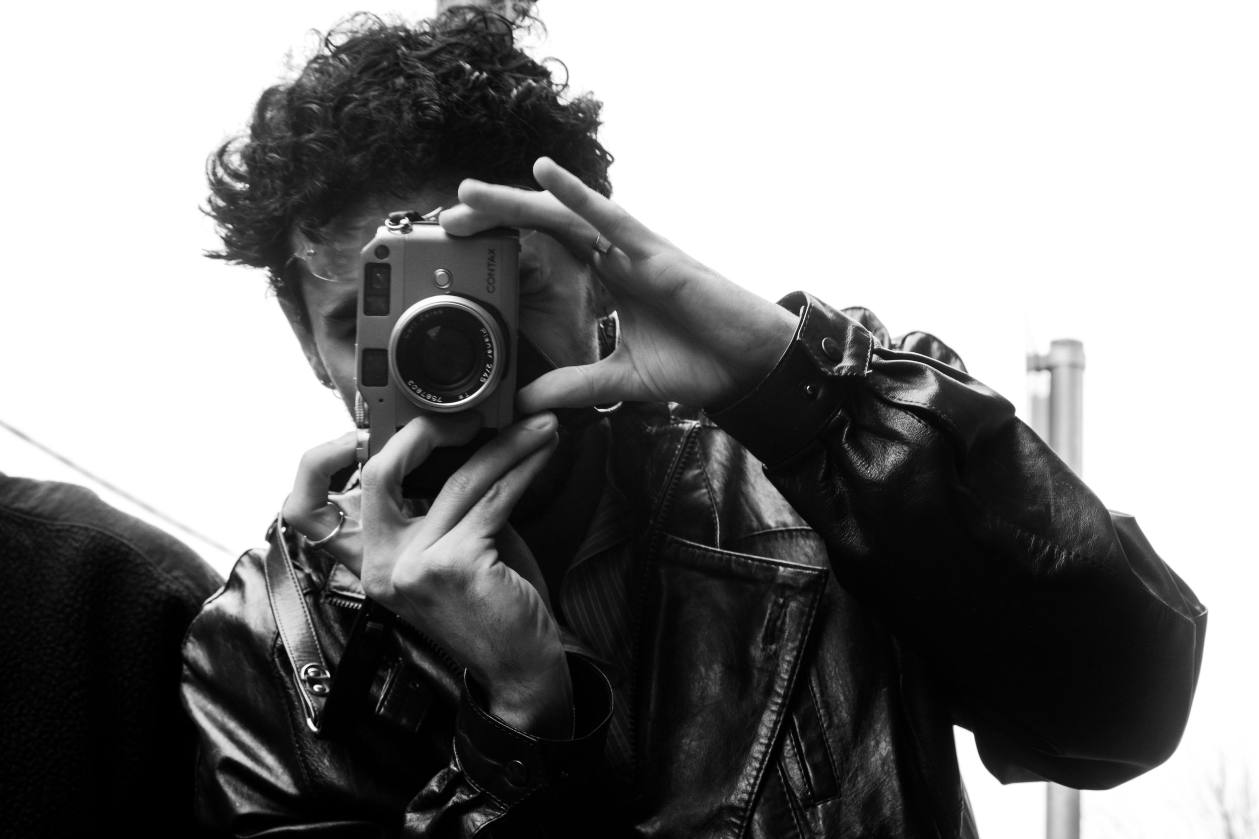 Person with curly hair wearing a leather jacket taking a photo with a camera.