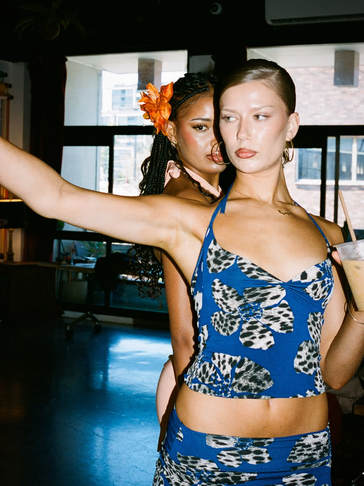 Two women standing indoors, one in front wearing a blue patterned tank top and the other behind her with an orange flower in her hair. The woman in front is holding a drink.