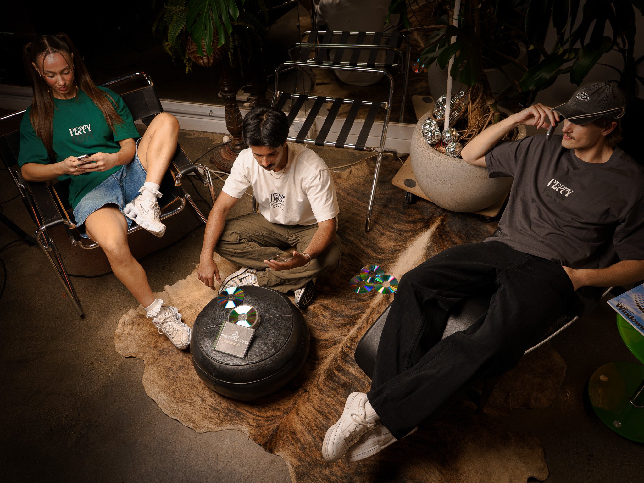 Three young people sitting in a casual indoor setting with plants and decorative items. One woman with long hair and a green T-shirt is using her phone, a man in a white T-shirt is sitting cross-legged with a phone, and another man in a dark T-shirt and cap is reclining in a chair. There are CDs and a book on an ottoman and rug, with a large gemstone-like decorative item nearby.