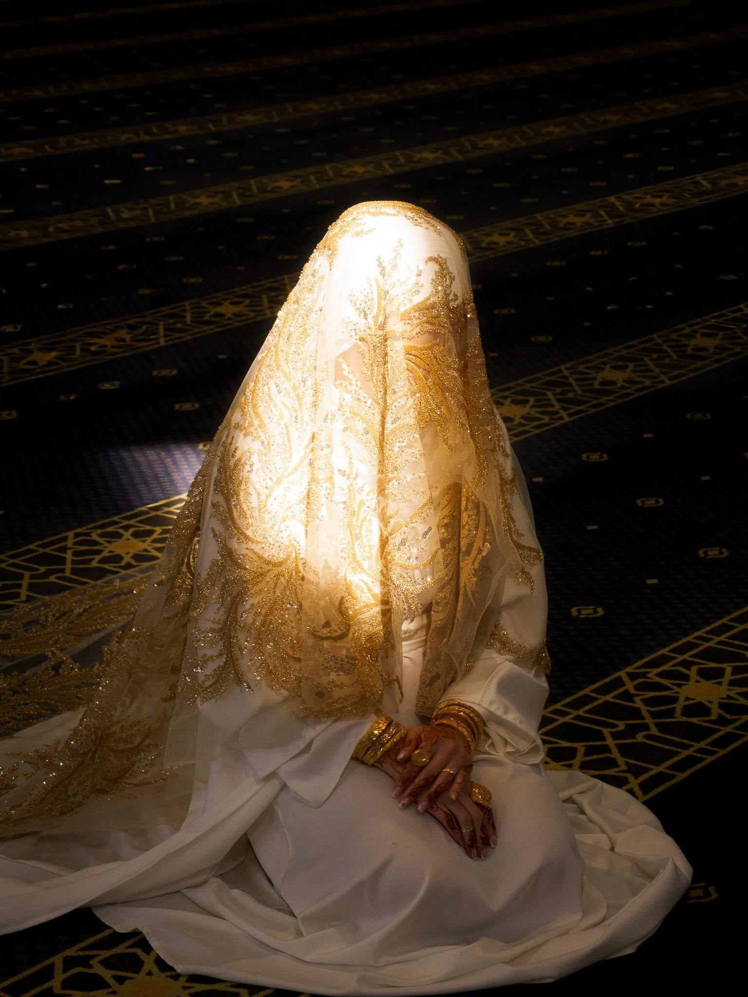 A woman dressed in white with gold embroidery covering her head and face, sitting on a patterned dark carpet with gold designs, illuminated by a beam of light.