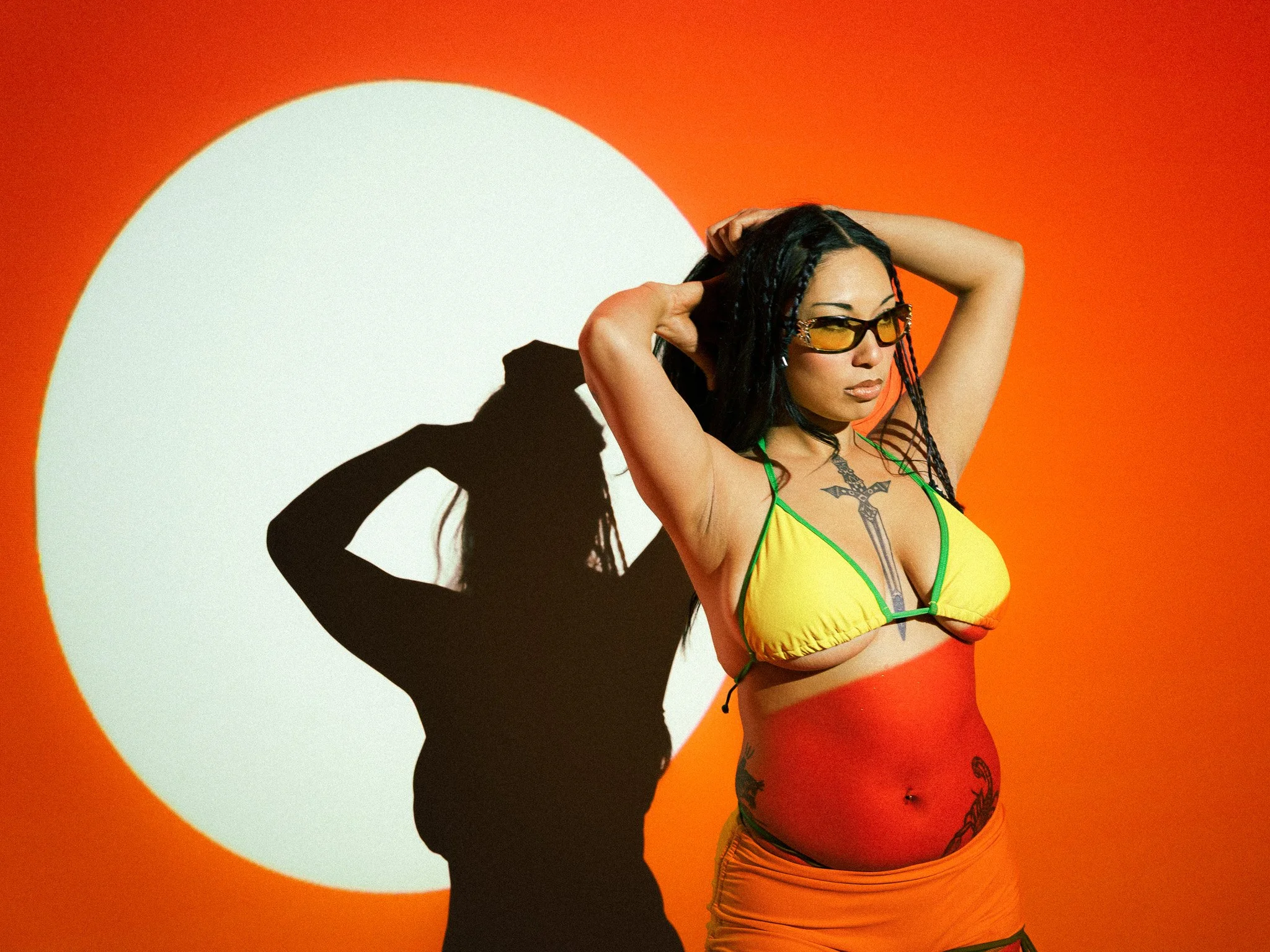 A woman with tattoos wearing sunglasses, a yellow bikini top, and orange shorts, standing against an orange background with a white circle shadow.