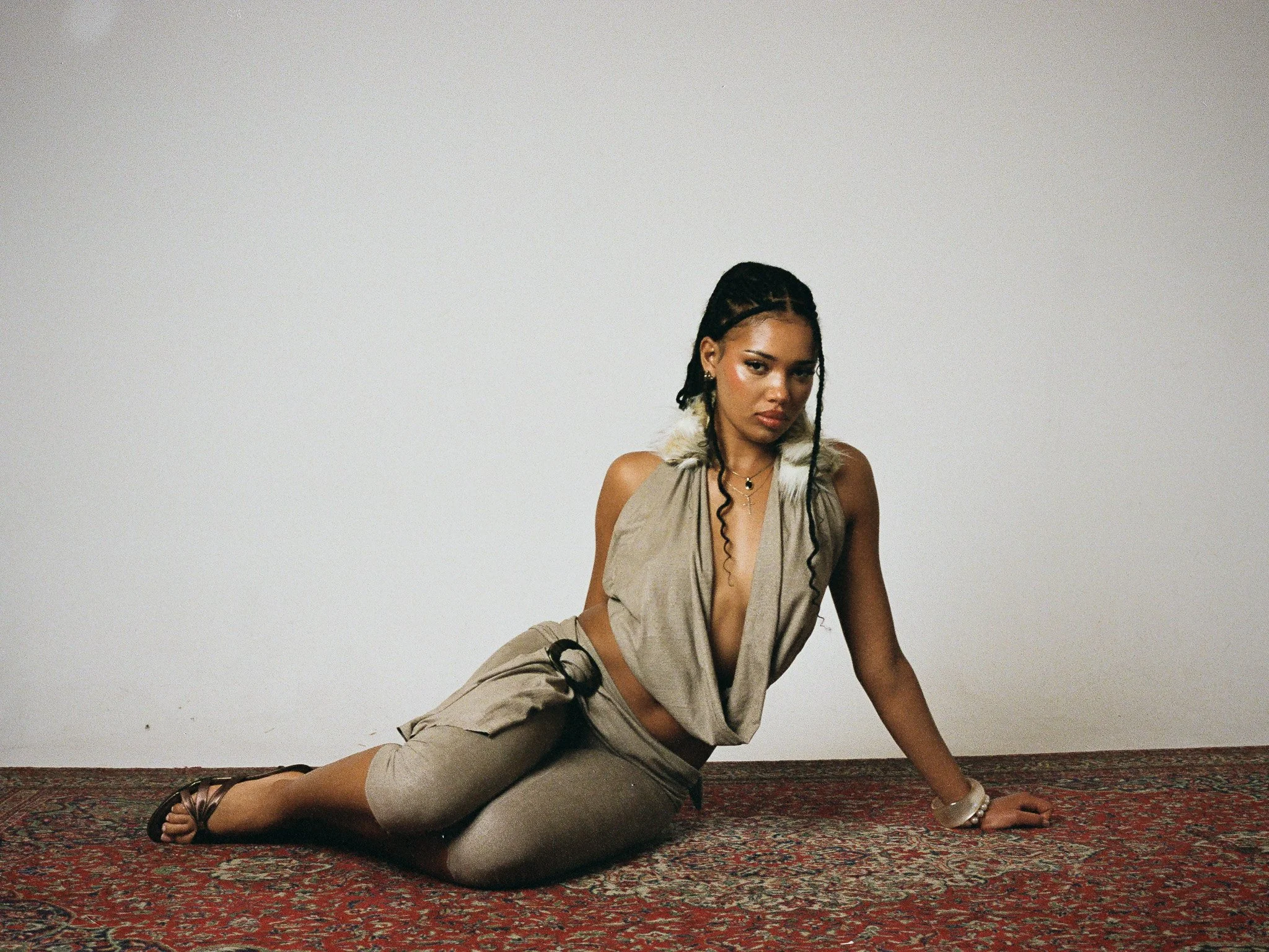 Young woman with dark hair styled in braids, dressed in beige clothing, sitting on a red patterned carpet against a plain white wall.