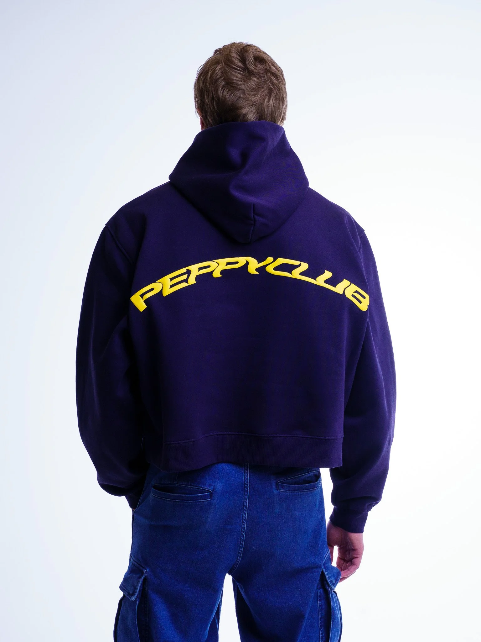 Back view of a person with brown hair wearing a navy hoodie with yellow text 'PEPPYCLUB' on the back, and blue jeans against a plain white background.