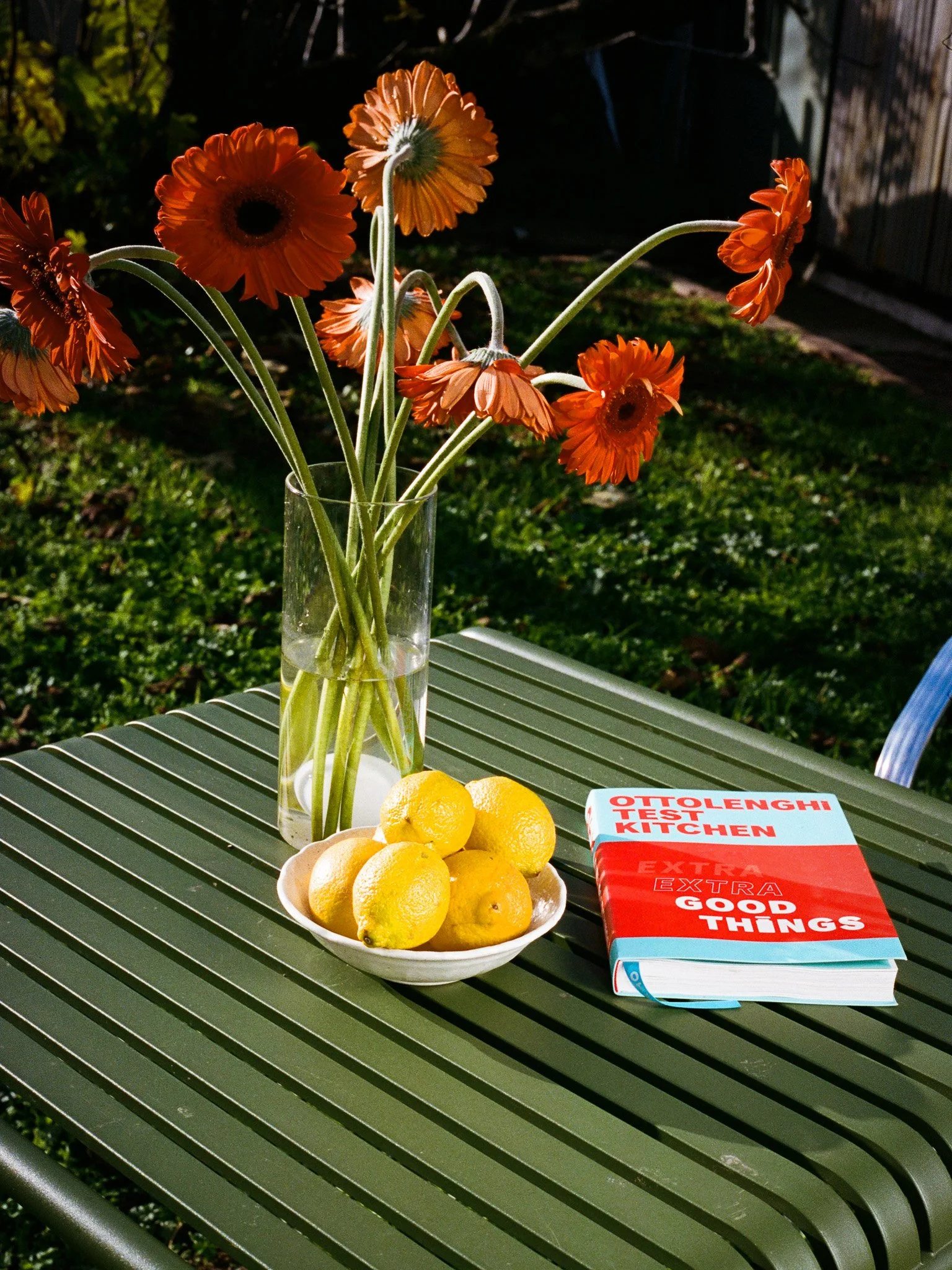 Bright orange gerbera daisies in a tall clear glass vase, a bowl of yellow lemons, and a red-and-white book titled 'Ottolenghi Test Kitchen: Extra Good Things' on a dark green outdoor table on a sunny day.