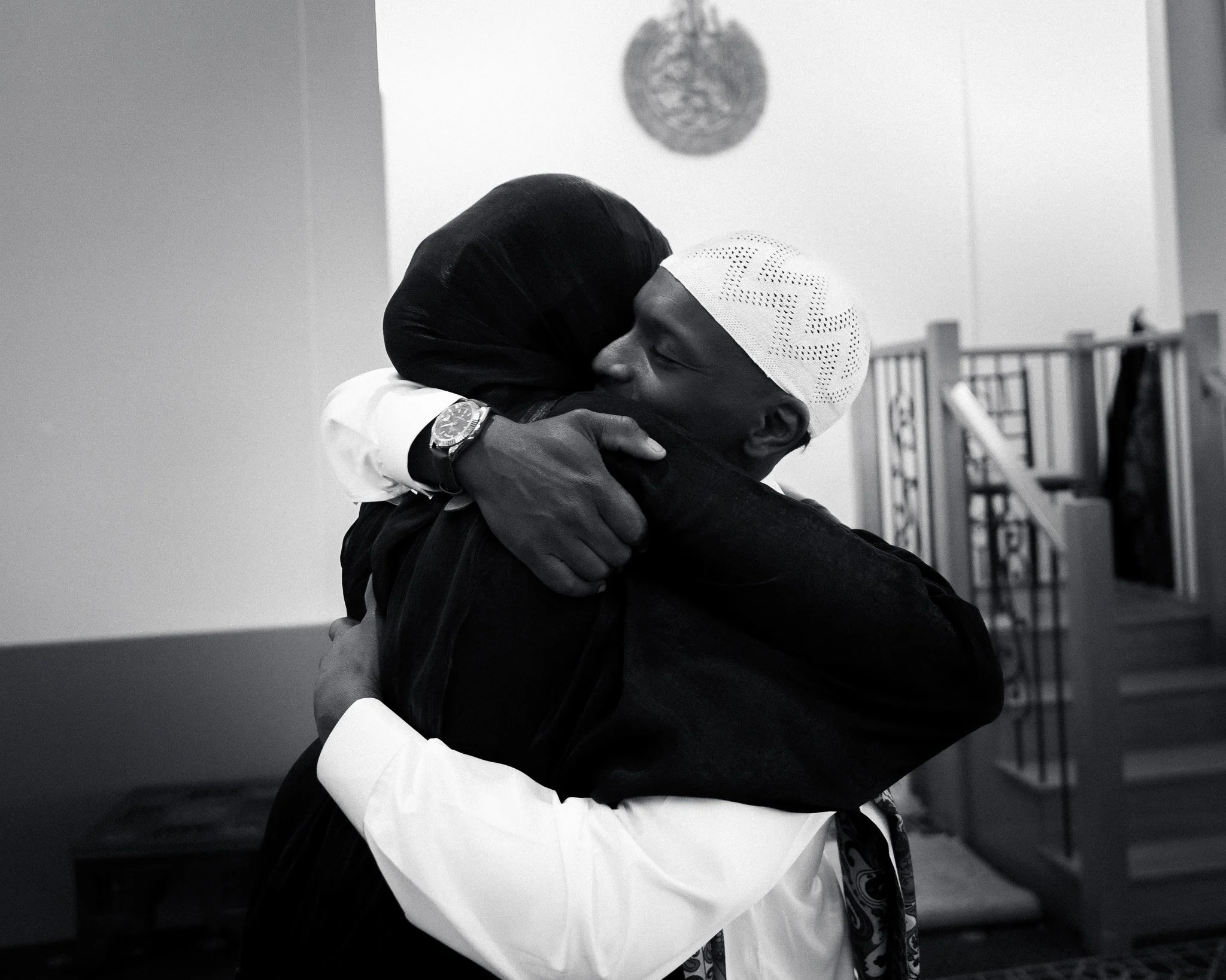 A young boy in traditional attire, wearing a white cap, embraces a woman dressed in black, inside a decorated room.