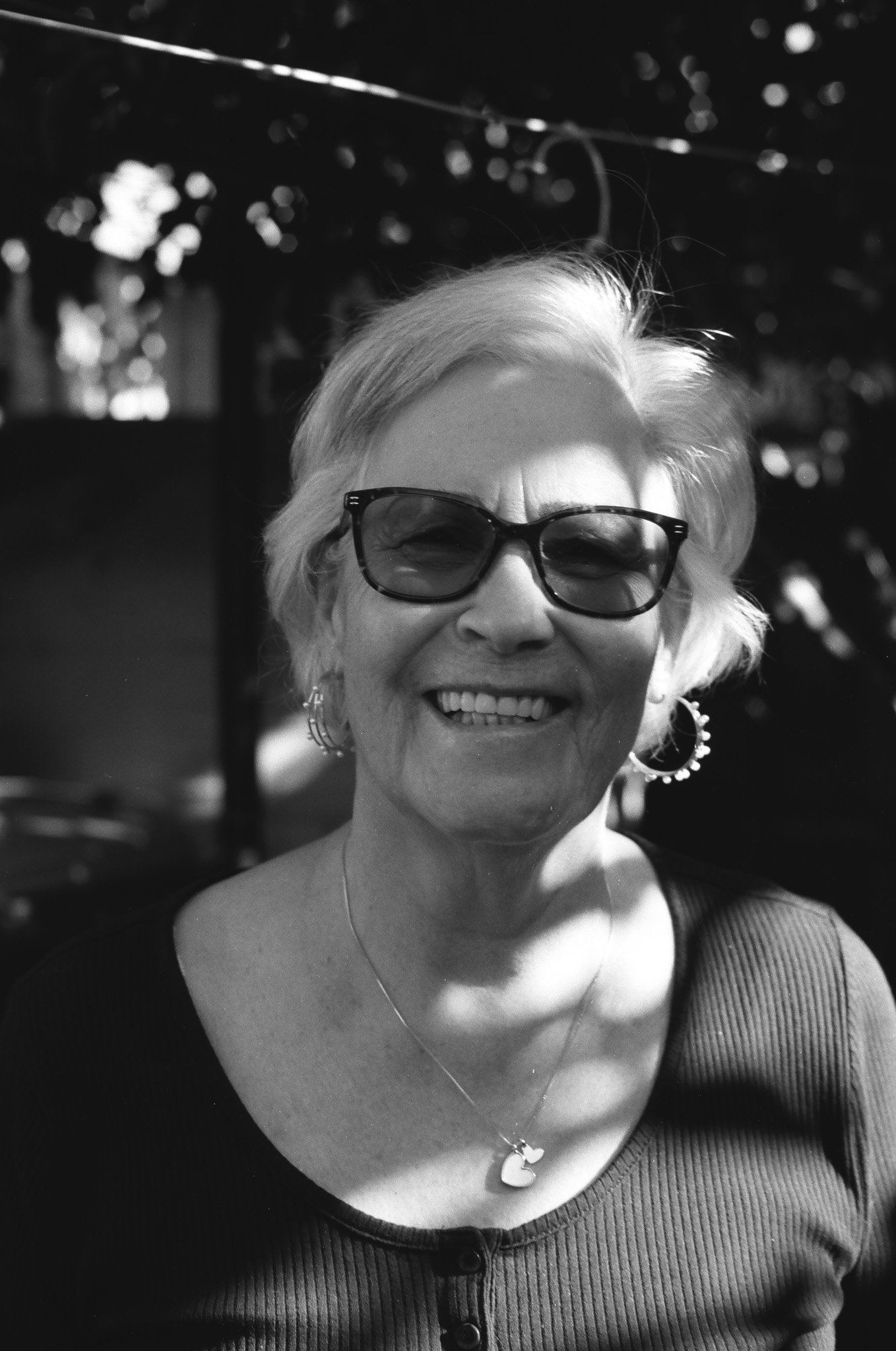 A smiling woman with short, wavy gray hair, wearing dark sunglasses, hoop earrings, a necklace with a moon-shaped pendant, and a black top, outdoors with sunlight and trees in the background.