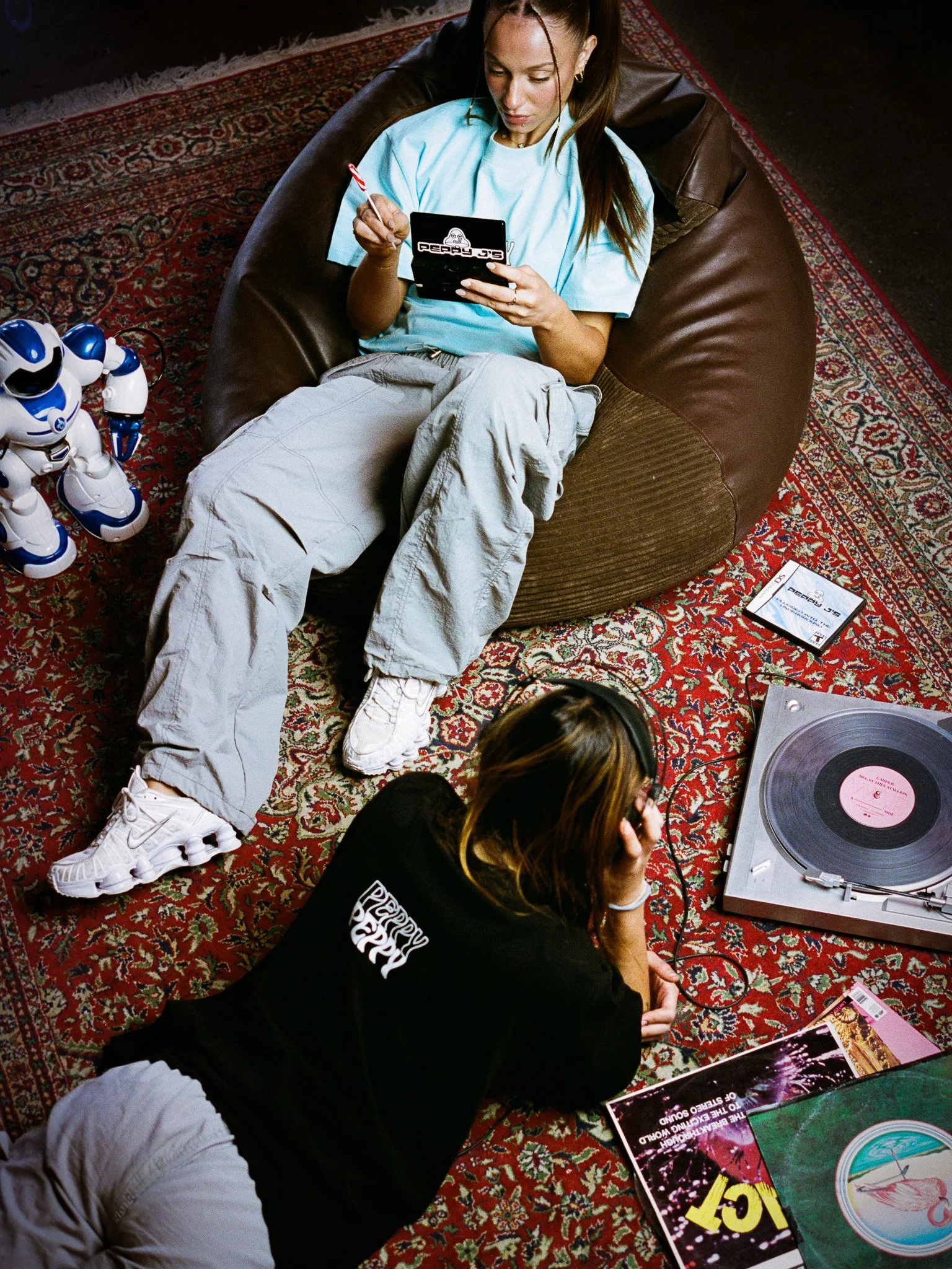A woman reclining on a bean bag chair, looking at her phone, with a toy robot nearby, and a woman lying on the floor next to a turntable, wearing headphones, surrounded by vinyl records.