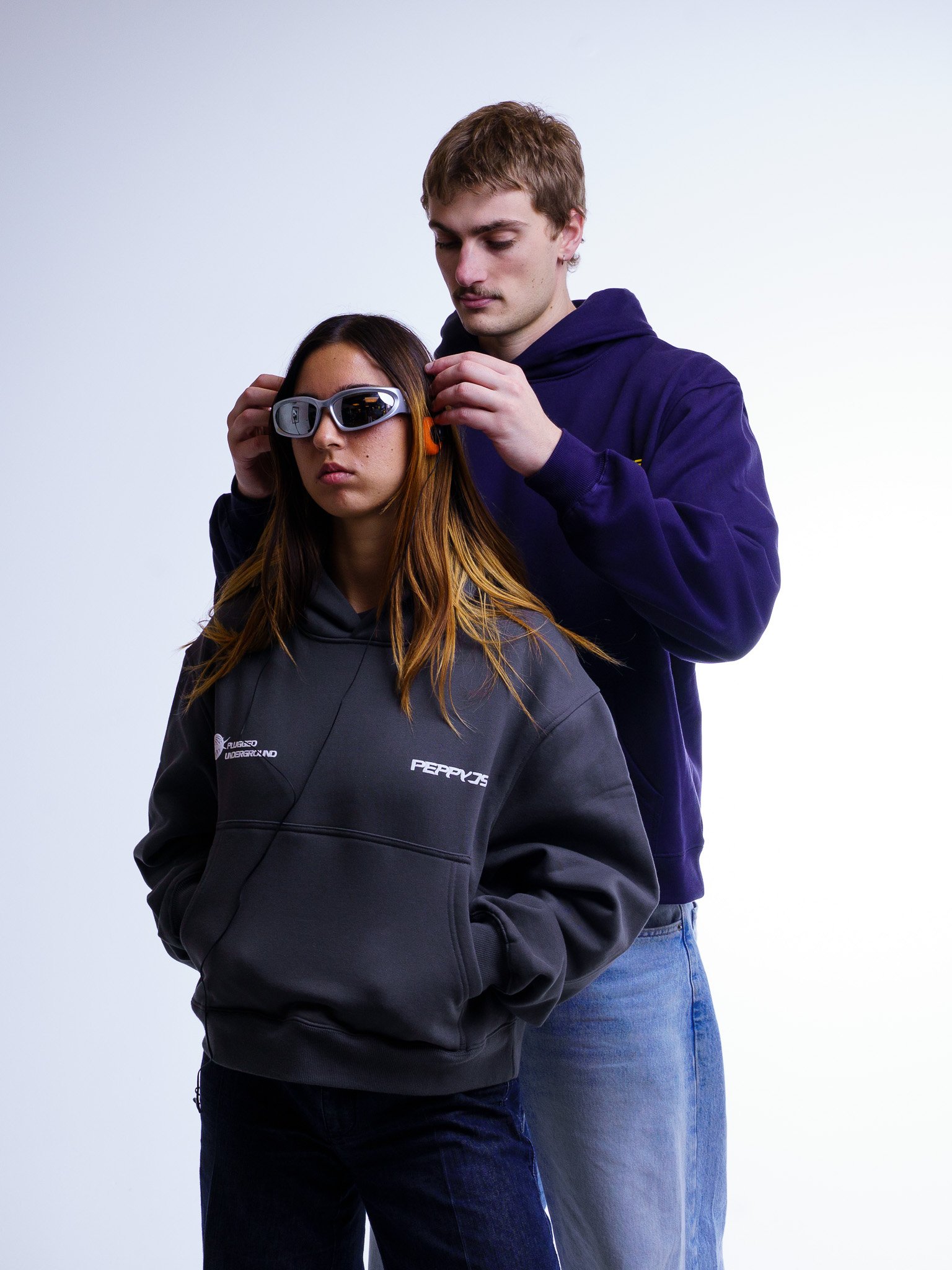 A young man helping a young woman try on glasses or goggles, with both wearing casual clothing and standing against a plain background.