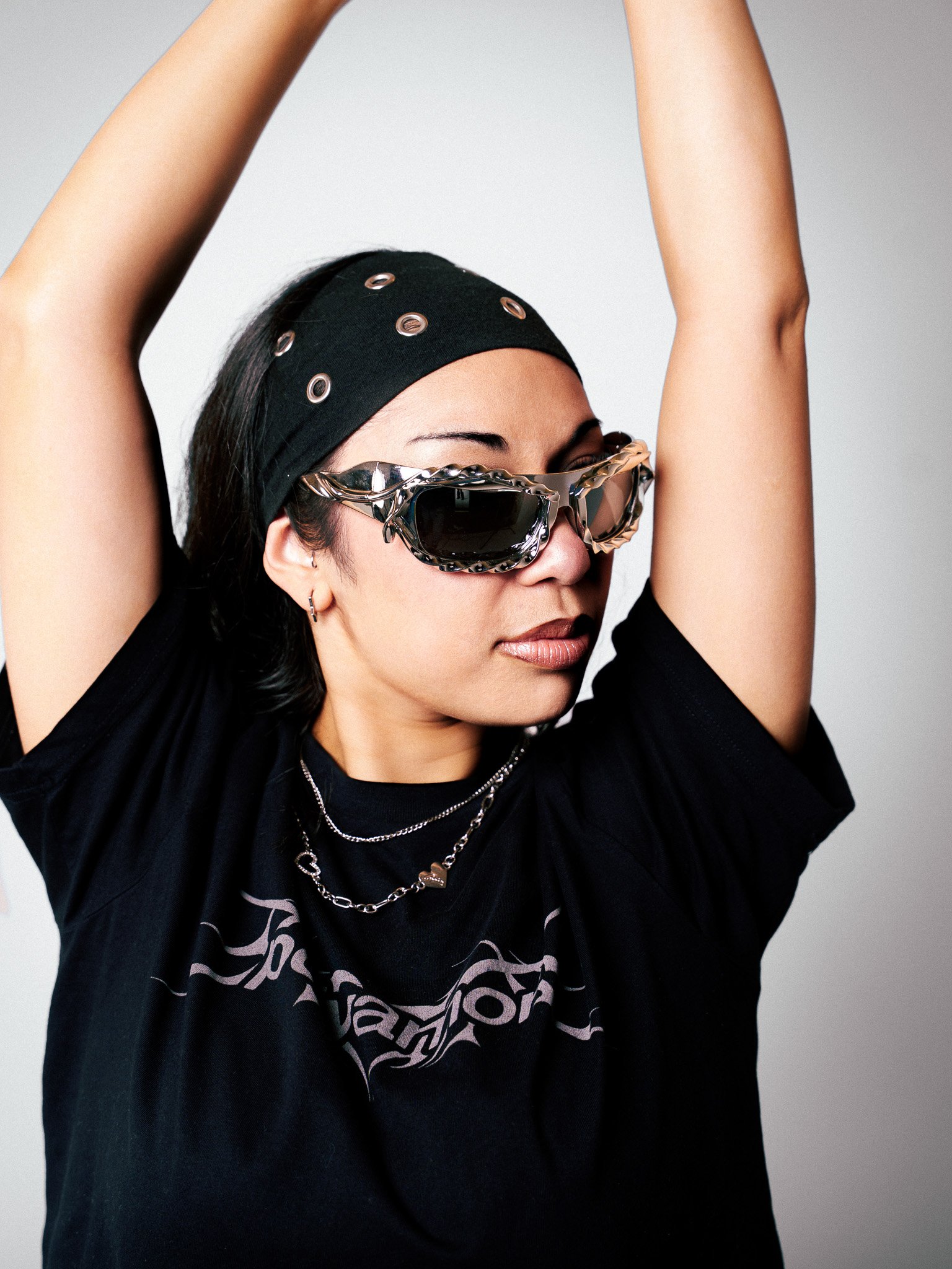 A woman with short dark hair in a black bandana, wearing patterned sunglasses, layered necklaces, and a black T-shirt, poses with her arms raised.