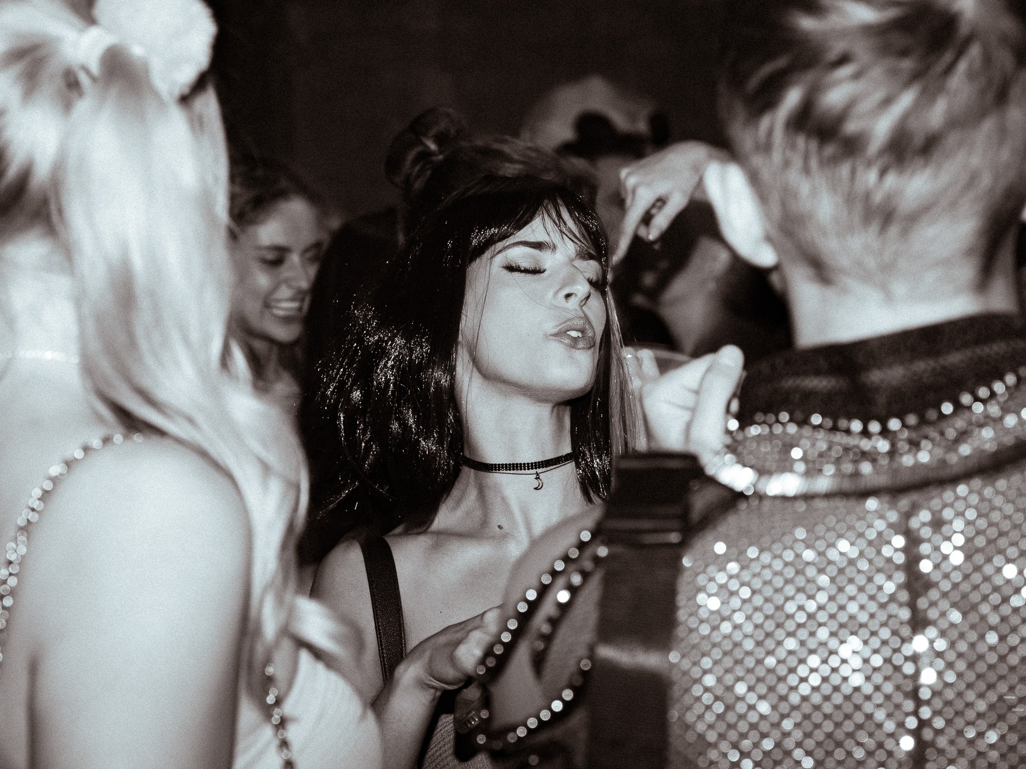 Group of people at a social gathering, with a woman with dark hair and a choker in the center, crowding around her.