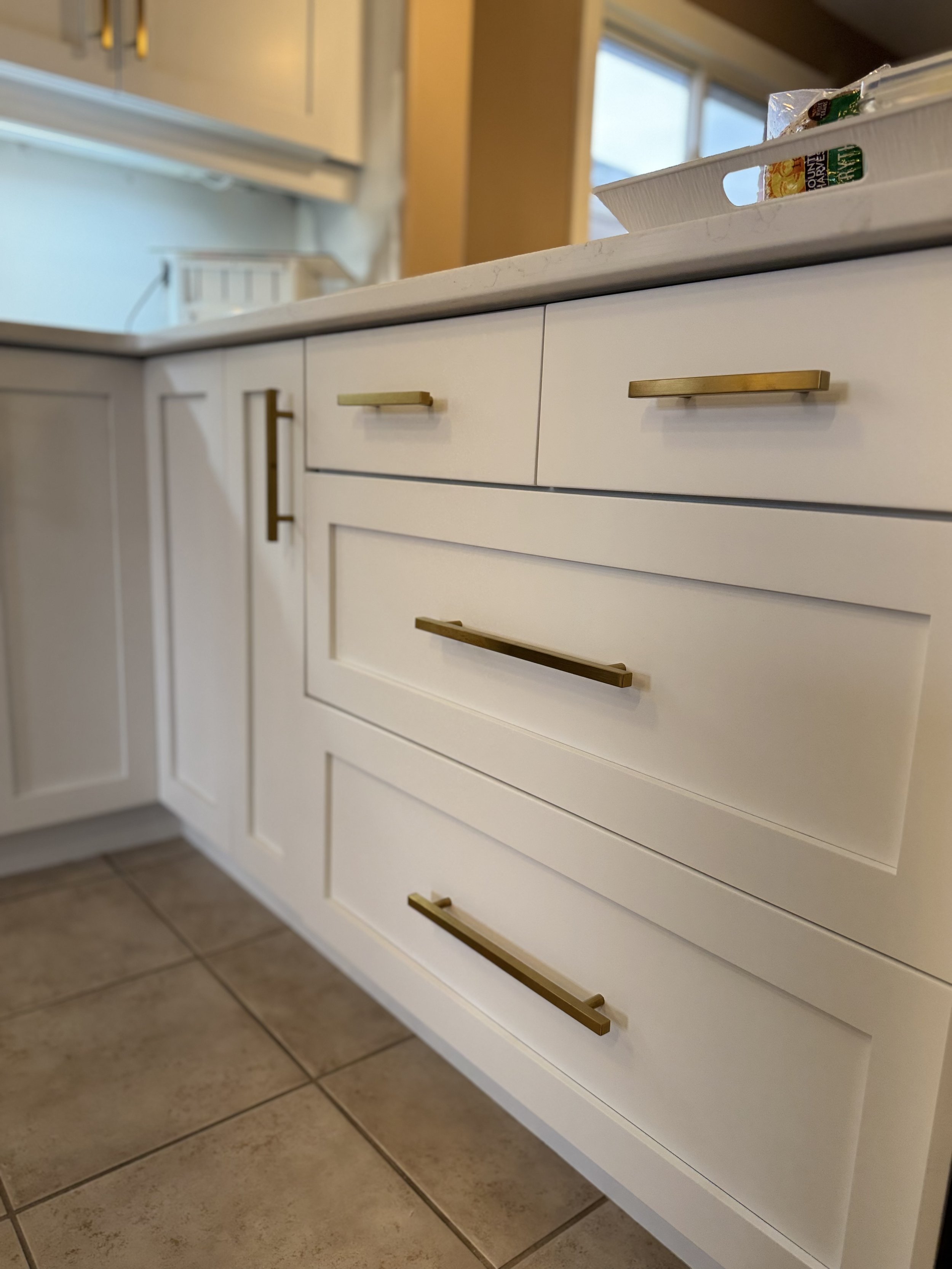 Close-up of white kitchen cabinets with gold handles and a beige tiled floor, with part of a countertop and a window in the background. Cabinet painting in Brentwood TN