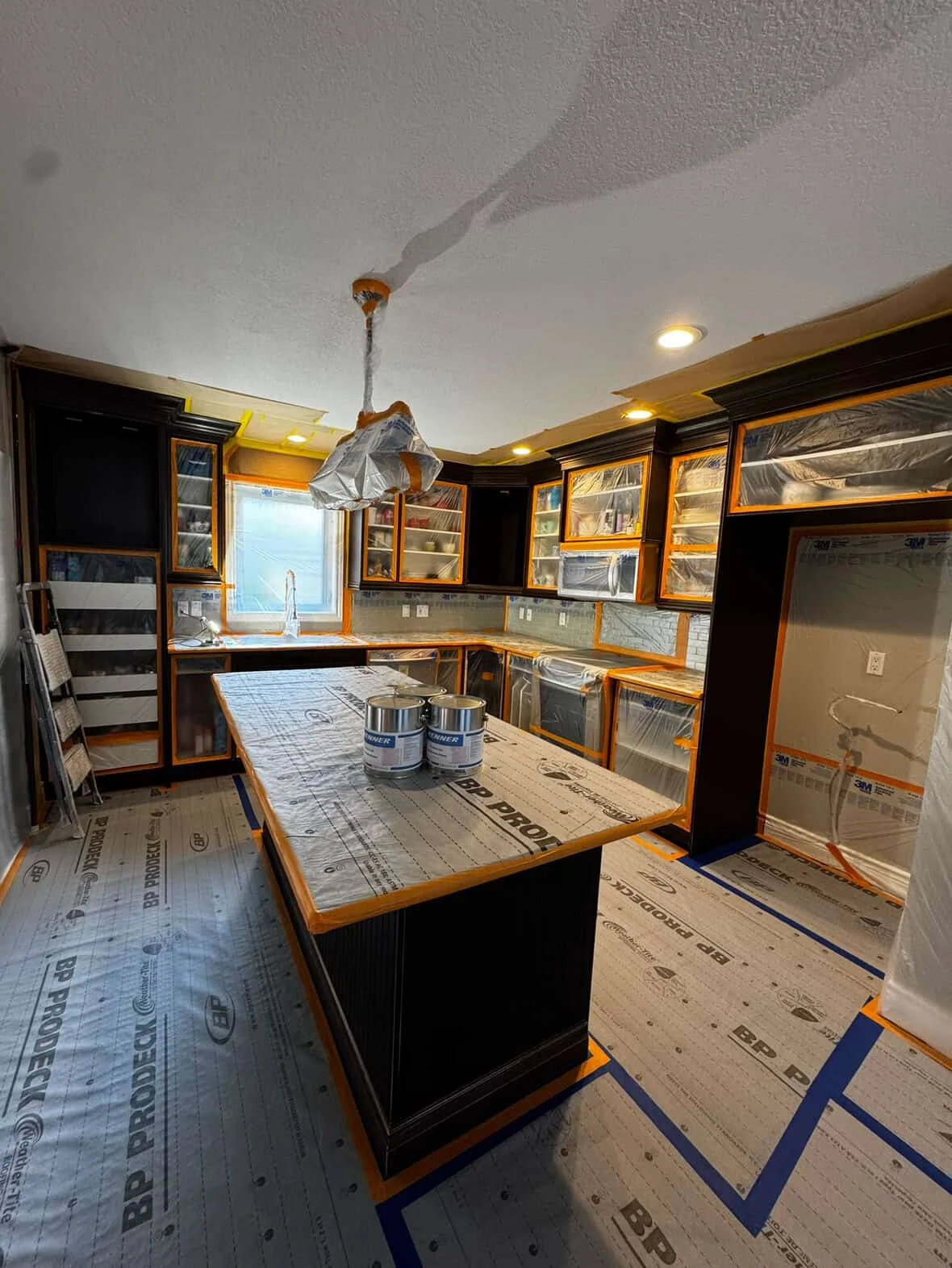 Kitchen under refinishing process with black cabinets, a large island, protected woodwork, and painting supplies.