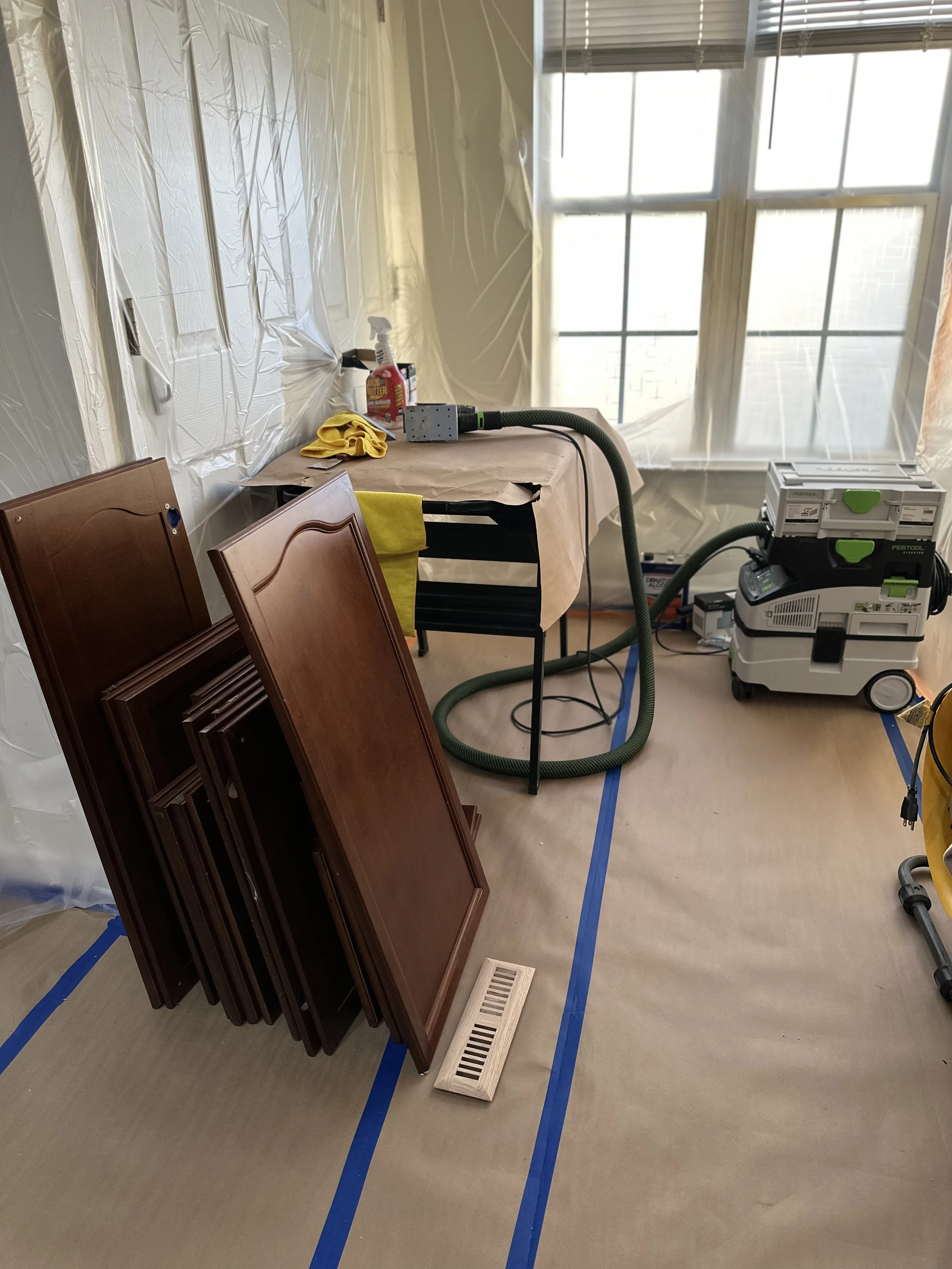 A partial view of a room in the process of cabinet refinishing with a table covered in painter's supplies, a vacuum, and stacked wooden cabinet doors.