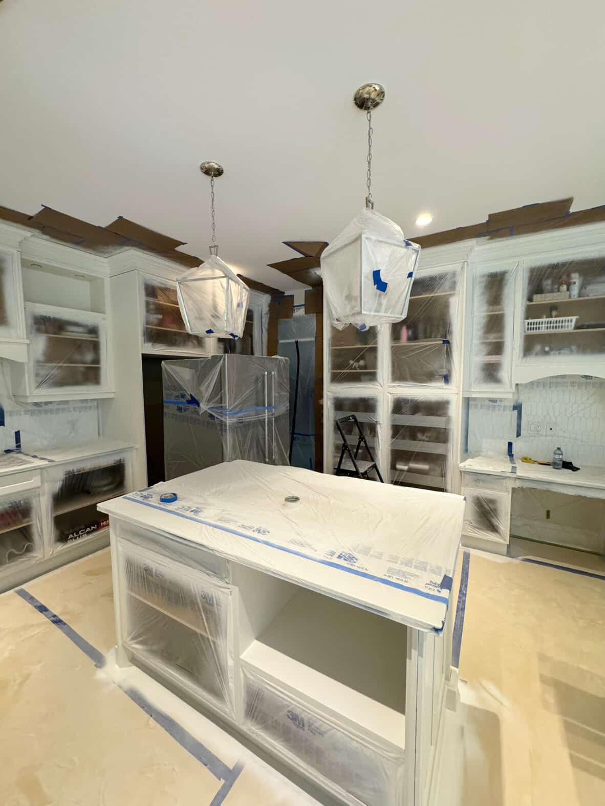 Kitchen undergoing refinishing process , with cabinets and island covered in painter's plastic, pendant lights covered in paper, and painter's tape on the floor.