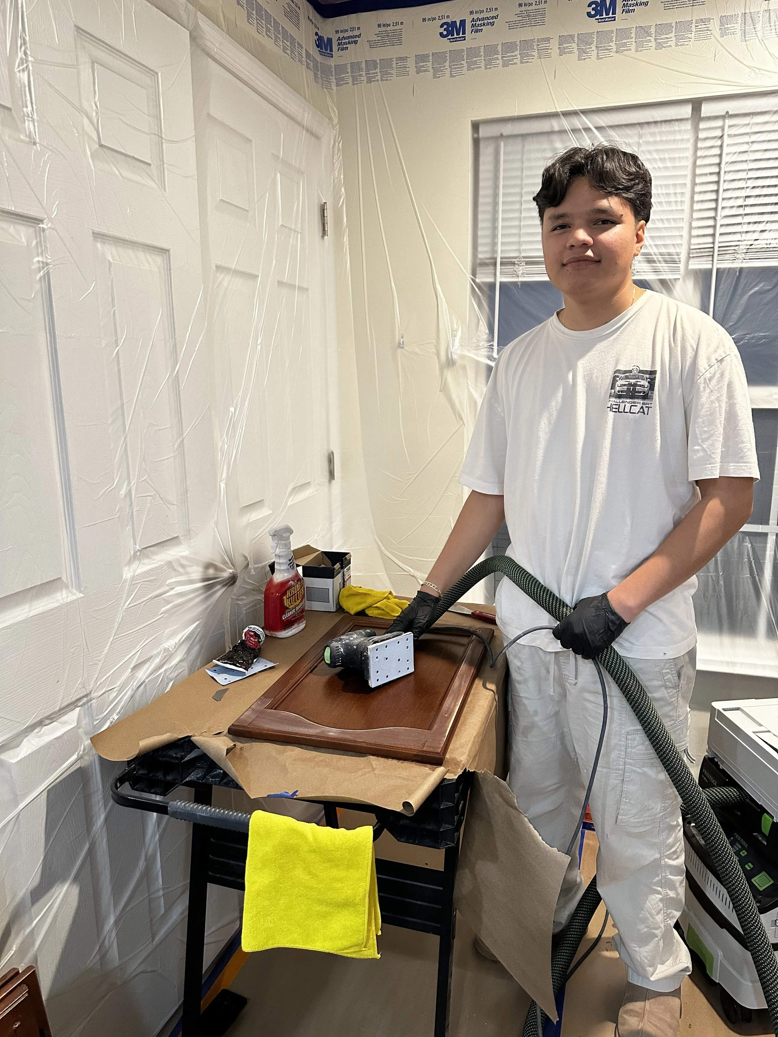 Young man in white t-shirt and gloves using a vacuum or spray device on a wooden cabinet, standing next to a cluttered workspace in a room with plastic sheeting covering the walls for painting or refinishing.