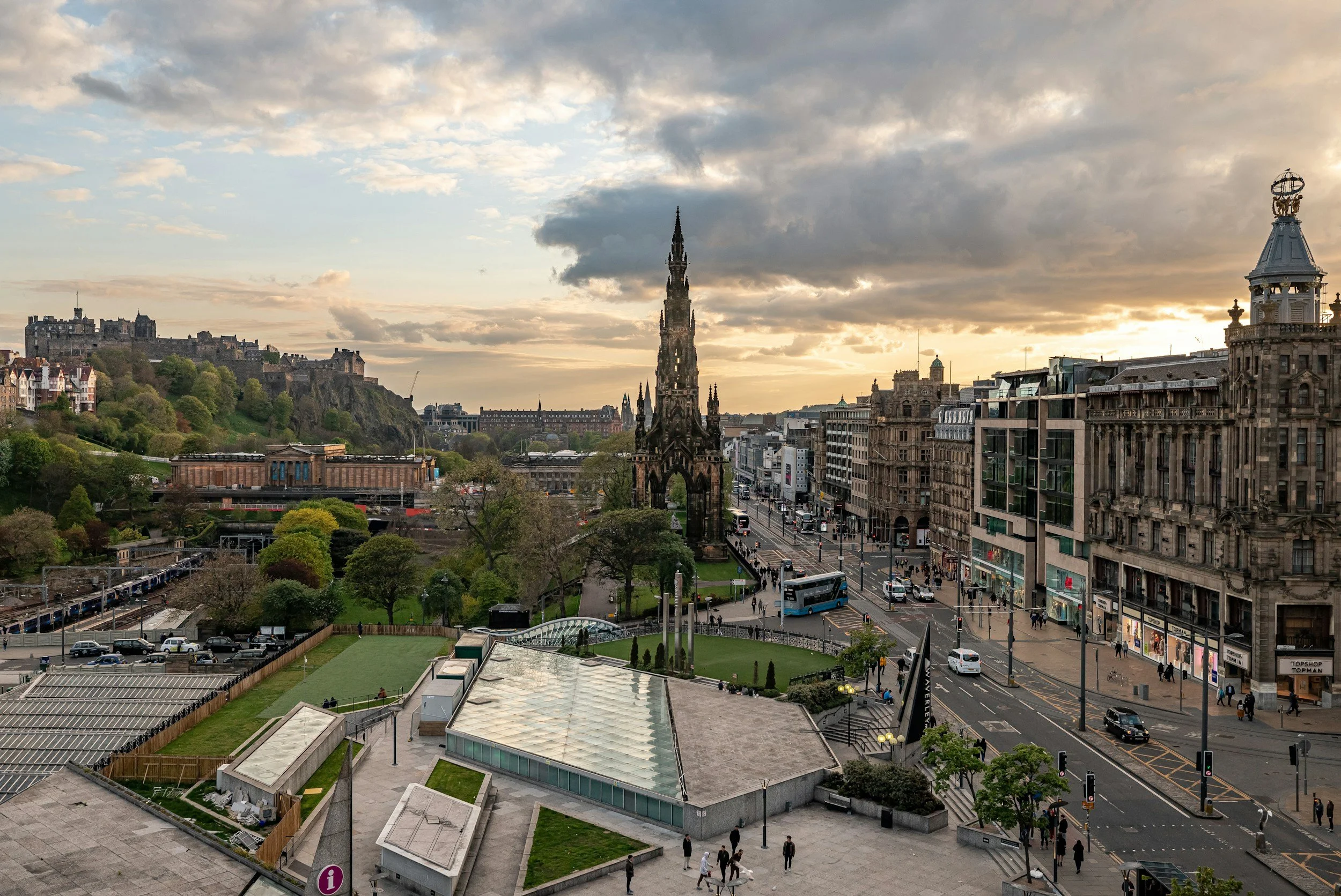 Edinburgh City Centre with historic and modern buildings, a gothic church spire, green park area, and a hilltop castle in the distance during cloudy sunset.