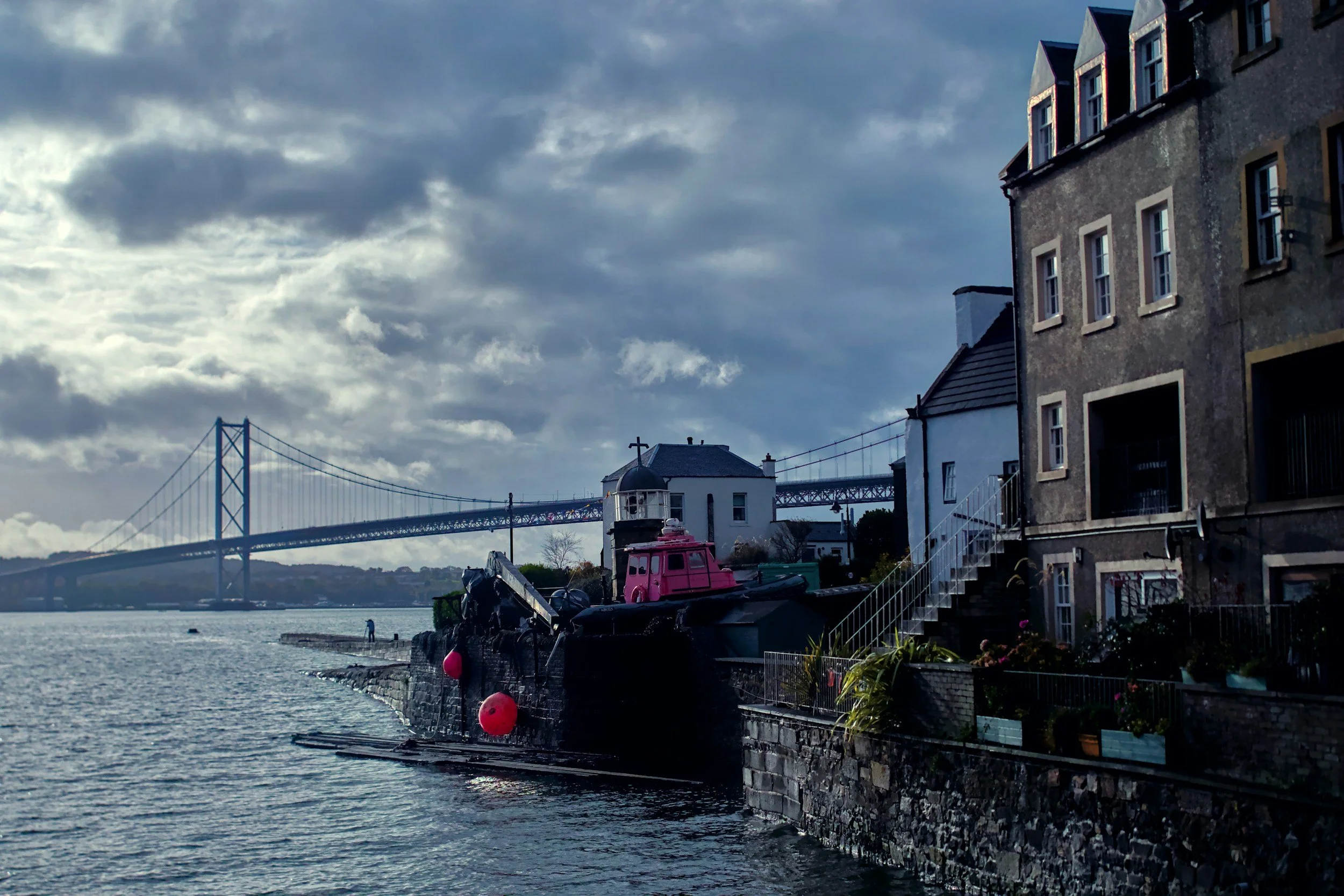 Scenic view of a waterfront with old buildings, a boat with a pink cabin, and a bridge in the background under a cloudy sky.
