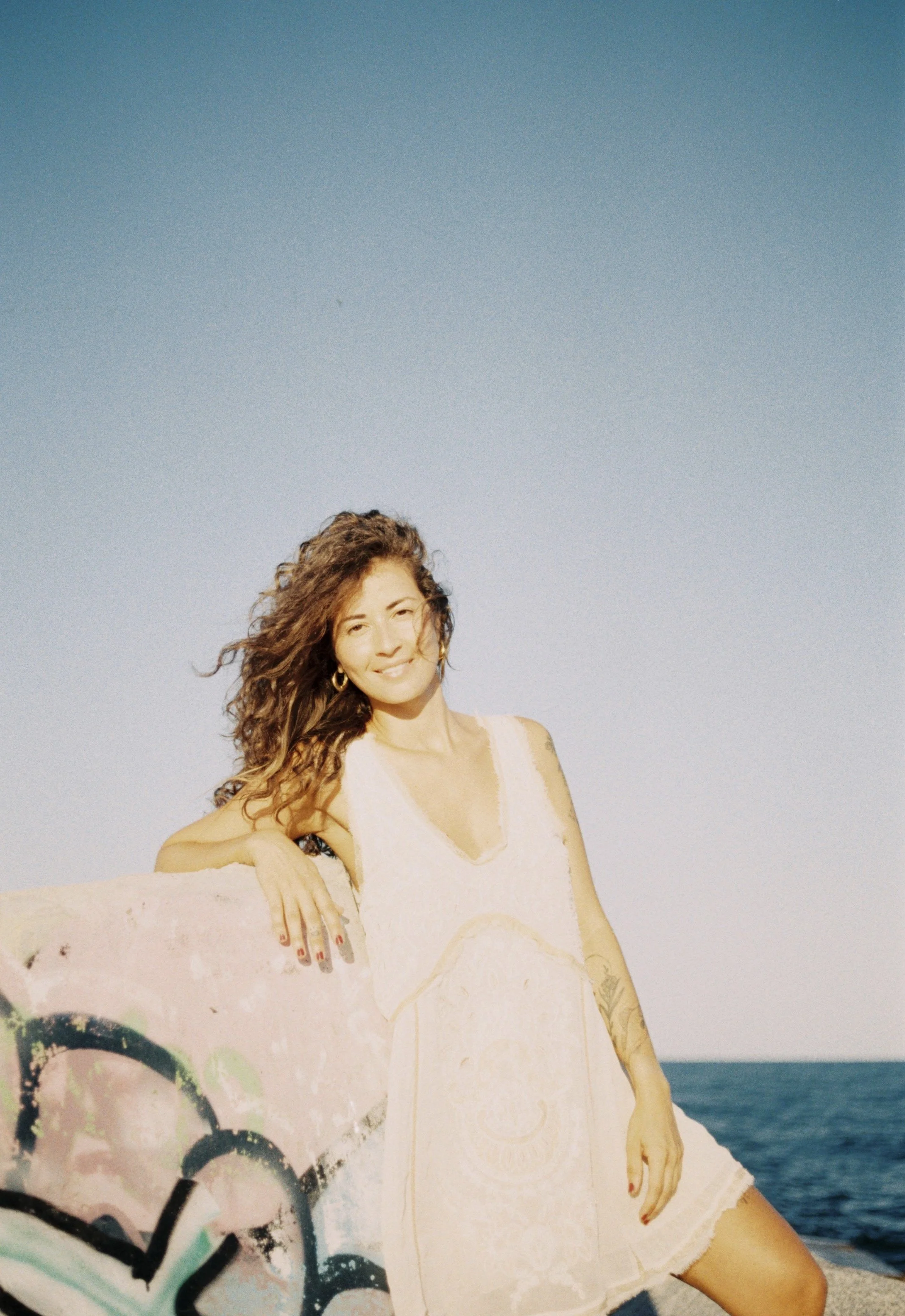 A woman with long curly hair standing outdoors near the water, smiling, wearing a white dress and gold hoop earrings, leaning against a graffiti-covered rock.