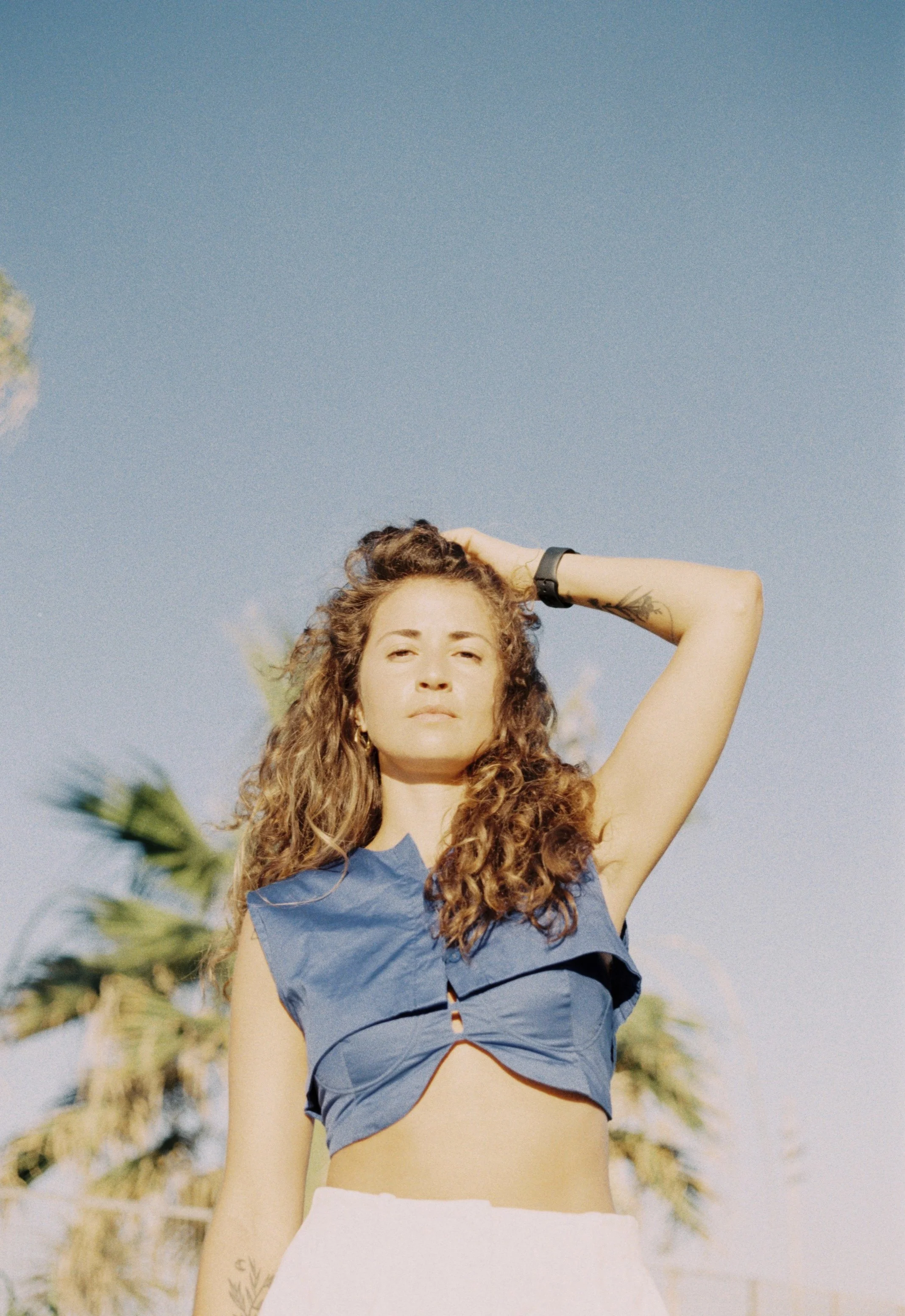 A woman standing outdoors with palm trees in the background, wearing a blue sleeveless top and white pants, with her arm raised to her head, gazing into the distance.