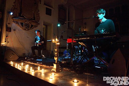 James Blake perform on stage with candles along the front edge. One plays a keyboard, the other plays an acoustic guitar. The scene is dimly lit with colored stage lights.