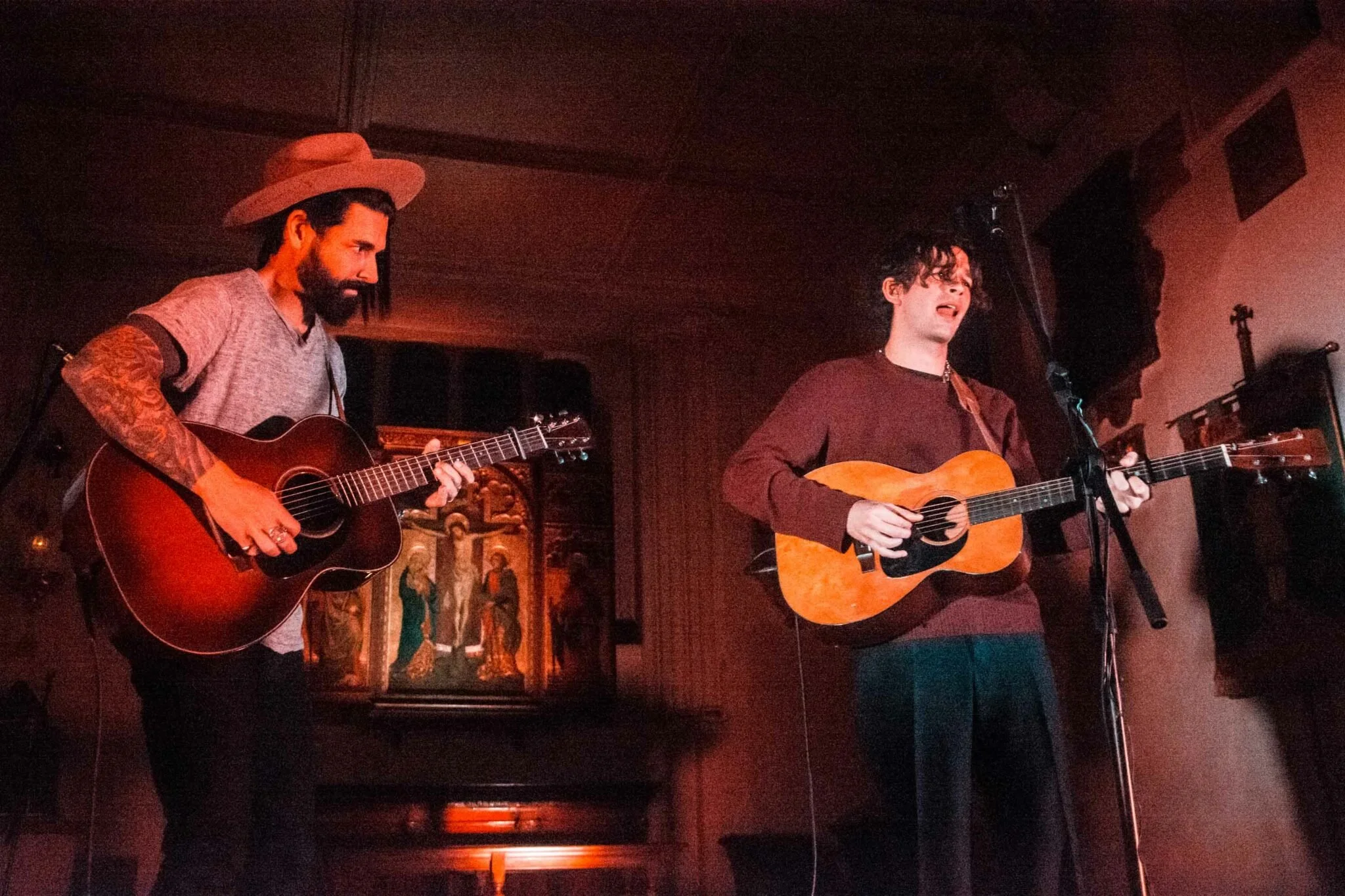 Dashboard Confessional and Matt Healy from the 1975 playing acoustic guitars on stage, one with tattoos and a hat, the other with curly hair, in an atmospheric room.