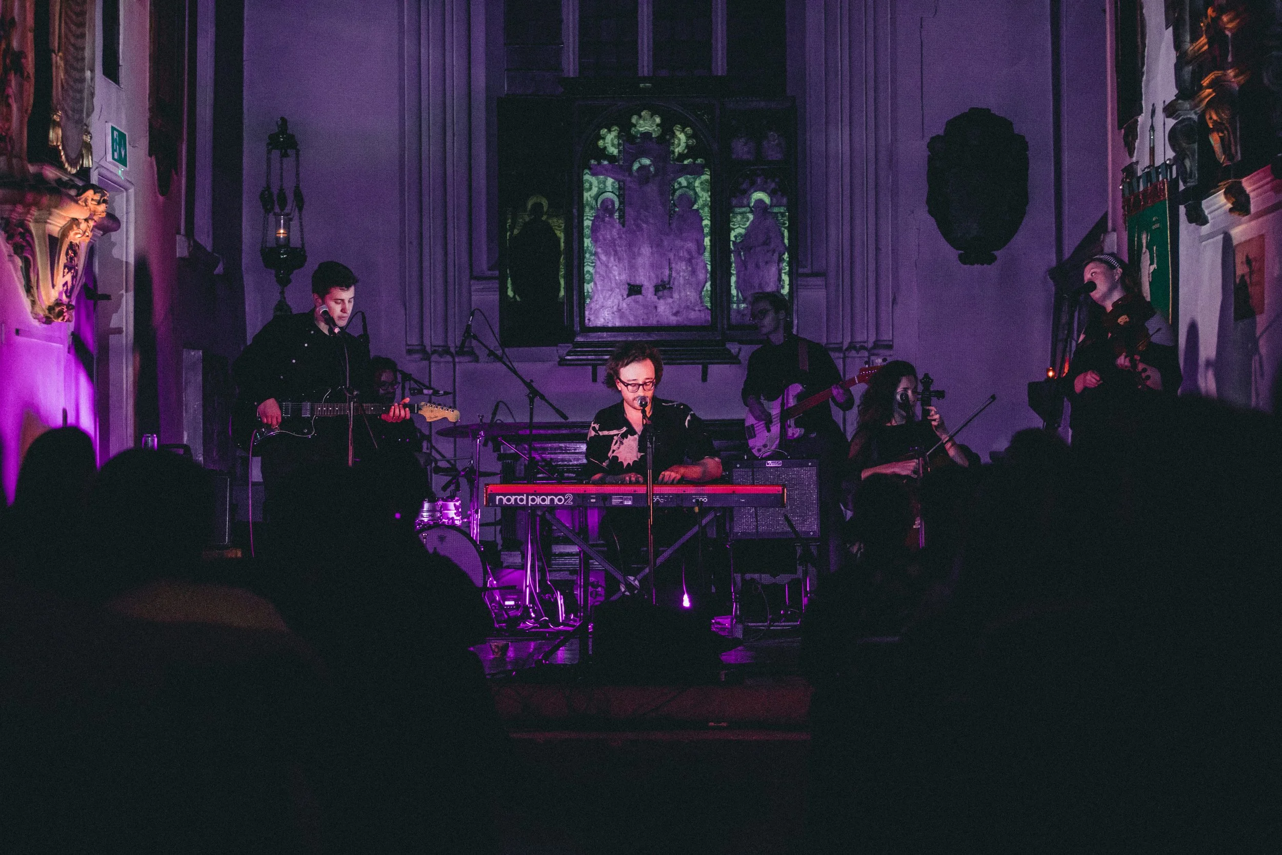 Wooden Arms performing on stage inside a church-like venue with purple lighting, including a keyboardist, guitarist, and violinist, with audience silhouettes in foreground.