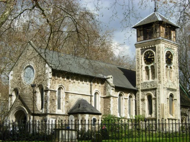 St Pancras Old Church, an old stone church with a tall clock tower, surrounded by a black wrought iron fence and trees.