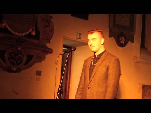 Sam Smith standing on stage in front of a microphone, wearing a brown blazer and jewelry, in a church with decorative wall art.