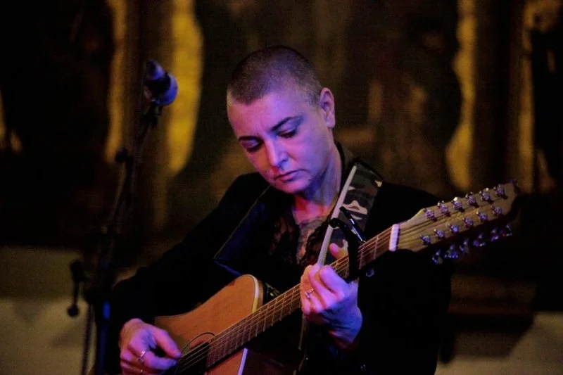 Sinead O Connor with short hair playing an acoustic guitar, performing on stage at night with dark background.