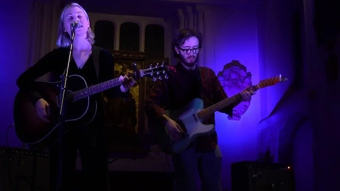 Phoebe Bridgers performs on stage, one singing with a guitar and the other playing an electric guitar, under purple lighting.