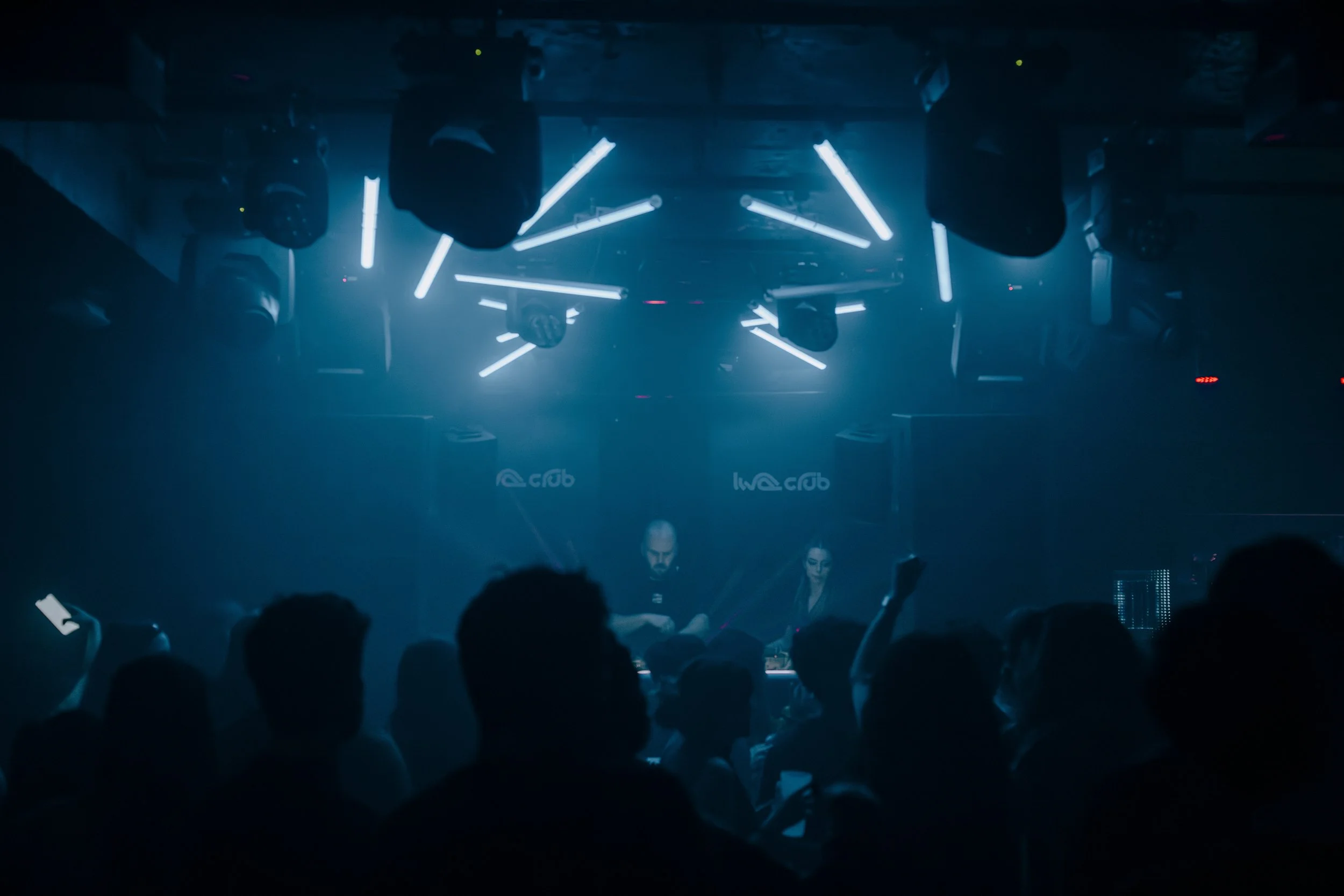 Une scène de club ou de concert avec des personnes dansant, éclairage bleu et plusieurs projecteurs suspendus au plafond.