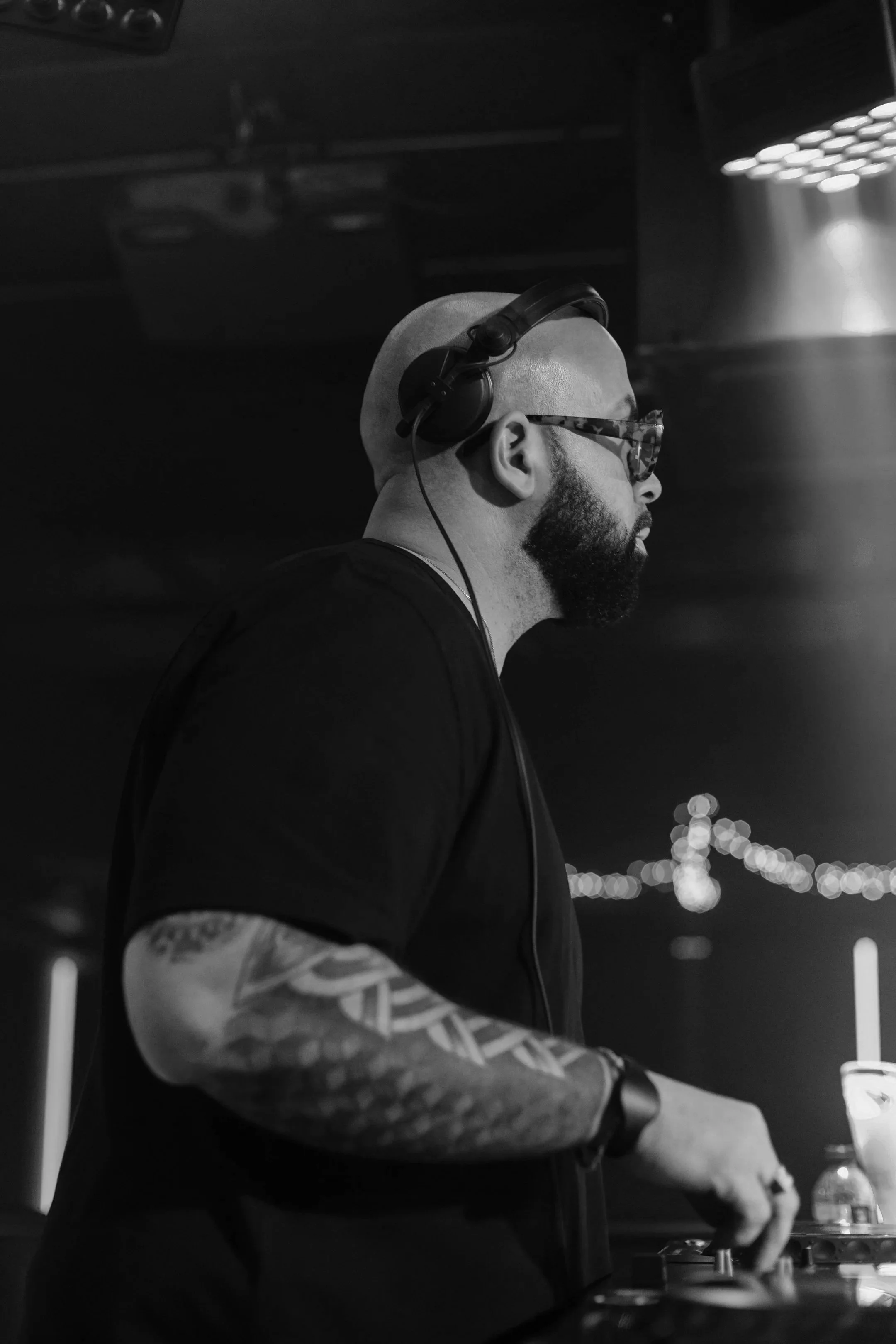 Un DJ avec des lunettes de soleil et une barbe, portant un casque audio et un t-shirt noir, en train de mixer lors d'une soirée, en noir et blanc.