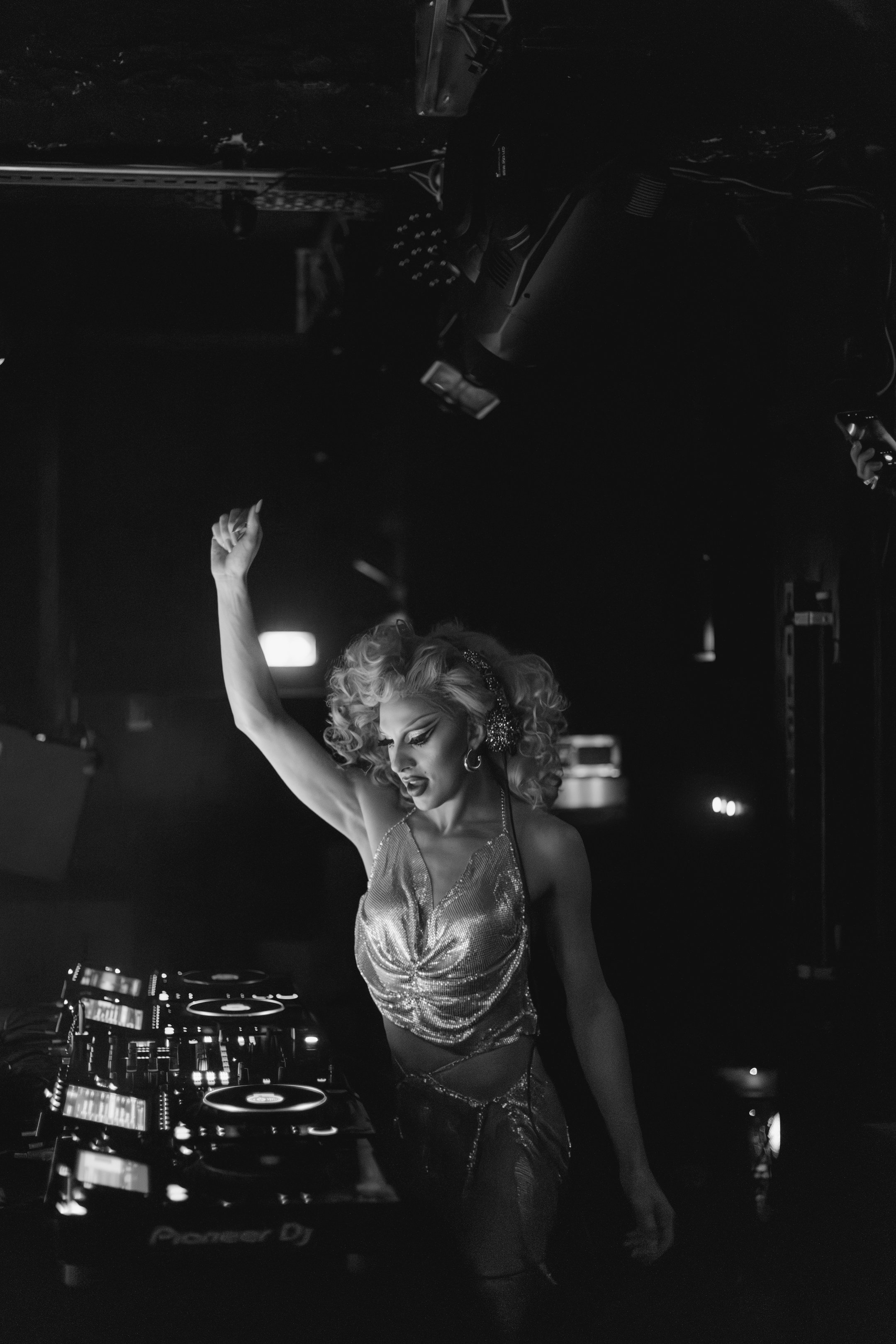 Une femme avec des cheveux bouclés, portant une tenue brillante, est DJ dans une boîte de nuit, levant une main en dansant.
