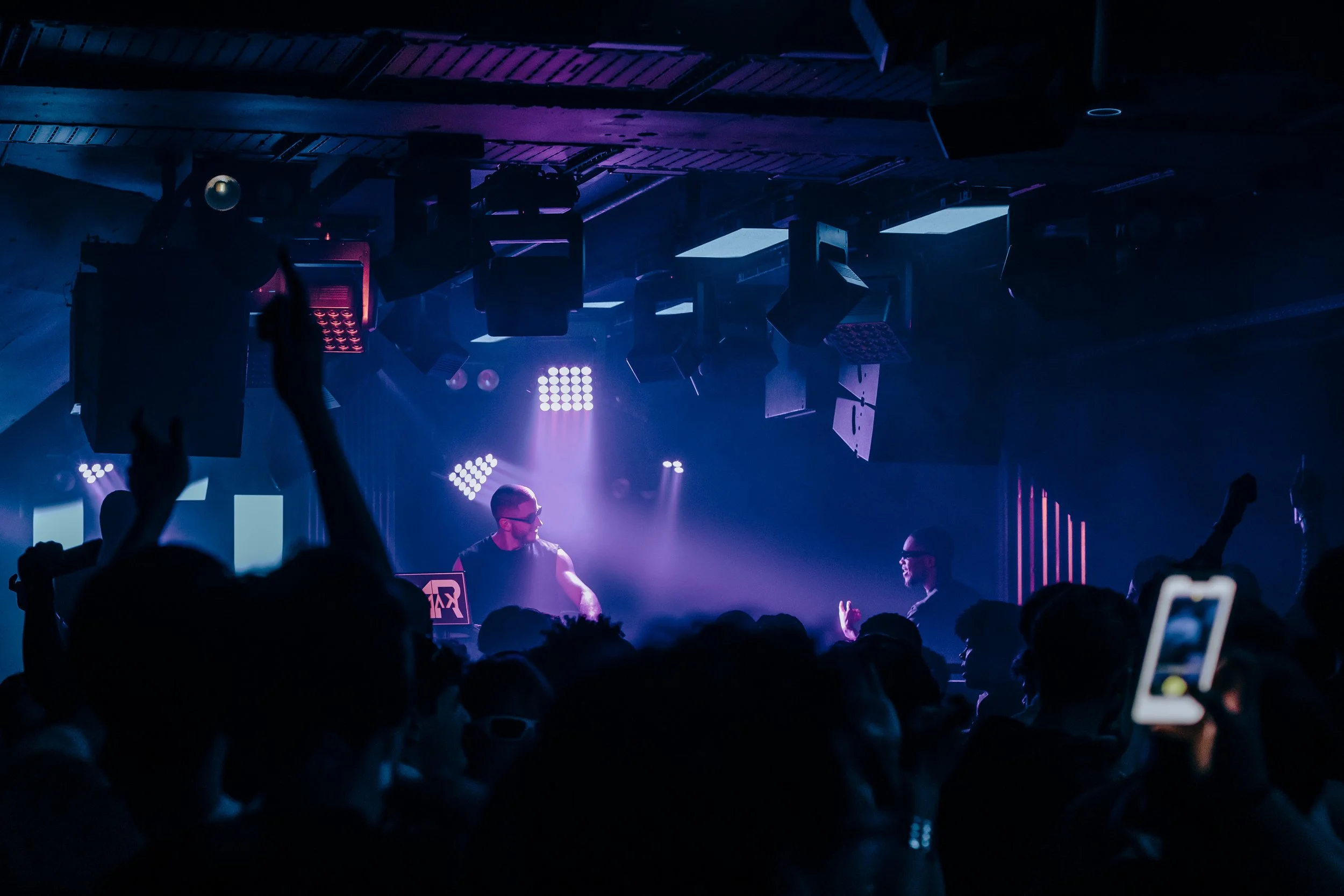 Une scène de concert ou de club avec deux DJ ou artistes sur scène, entourés de lumière bleue et violette, et une foule de spectateurs devant eux, certains levant la main ou prenant des photos avec leur téléphone.