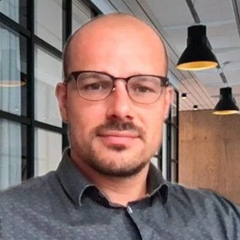 A man with glasses and a goatee, wearing a dark collared shirt, standing inside a modern office space.
