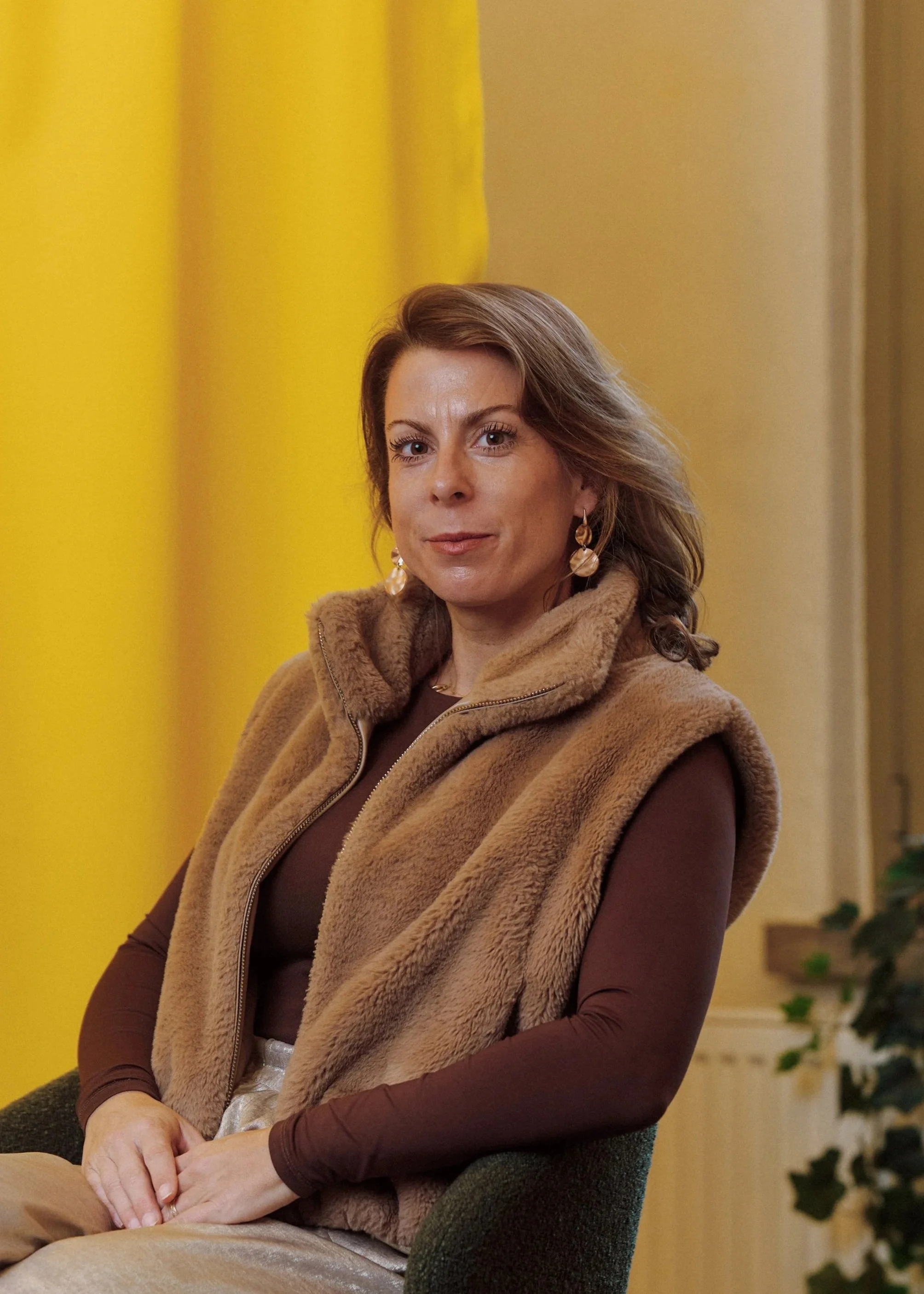 A woman with light brown, wavy hair and earrings, wearing a brown long-sleeve top and a tan furry vest, sitting on a dark green chair in front of yellow curtains.