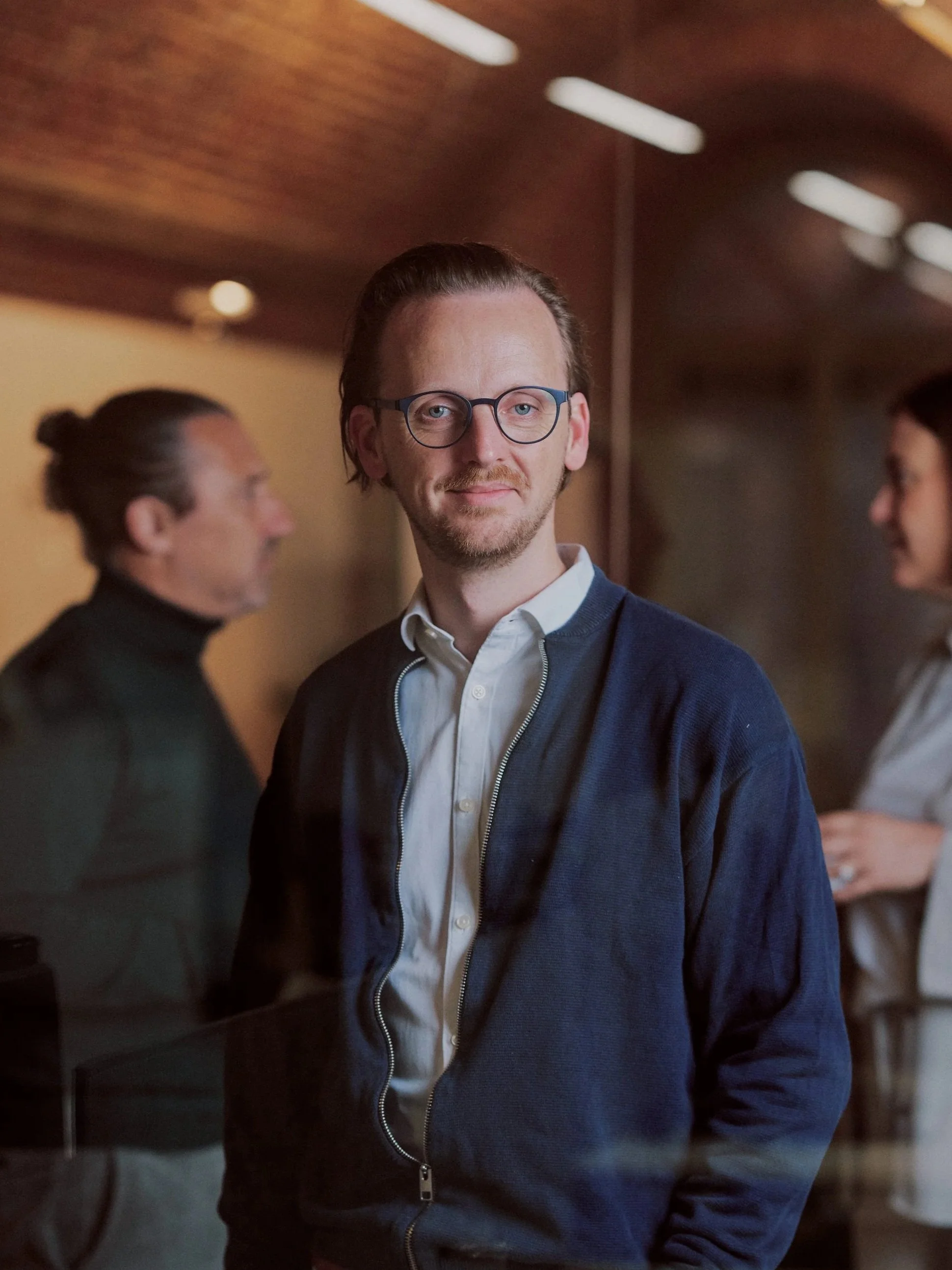 A man with glasses and a beard looking toward the camera while standing indoors, blurred people in the background.