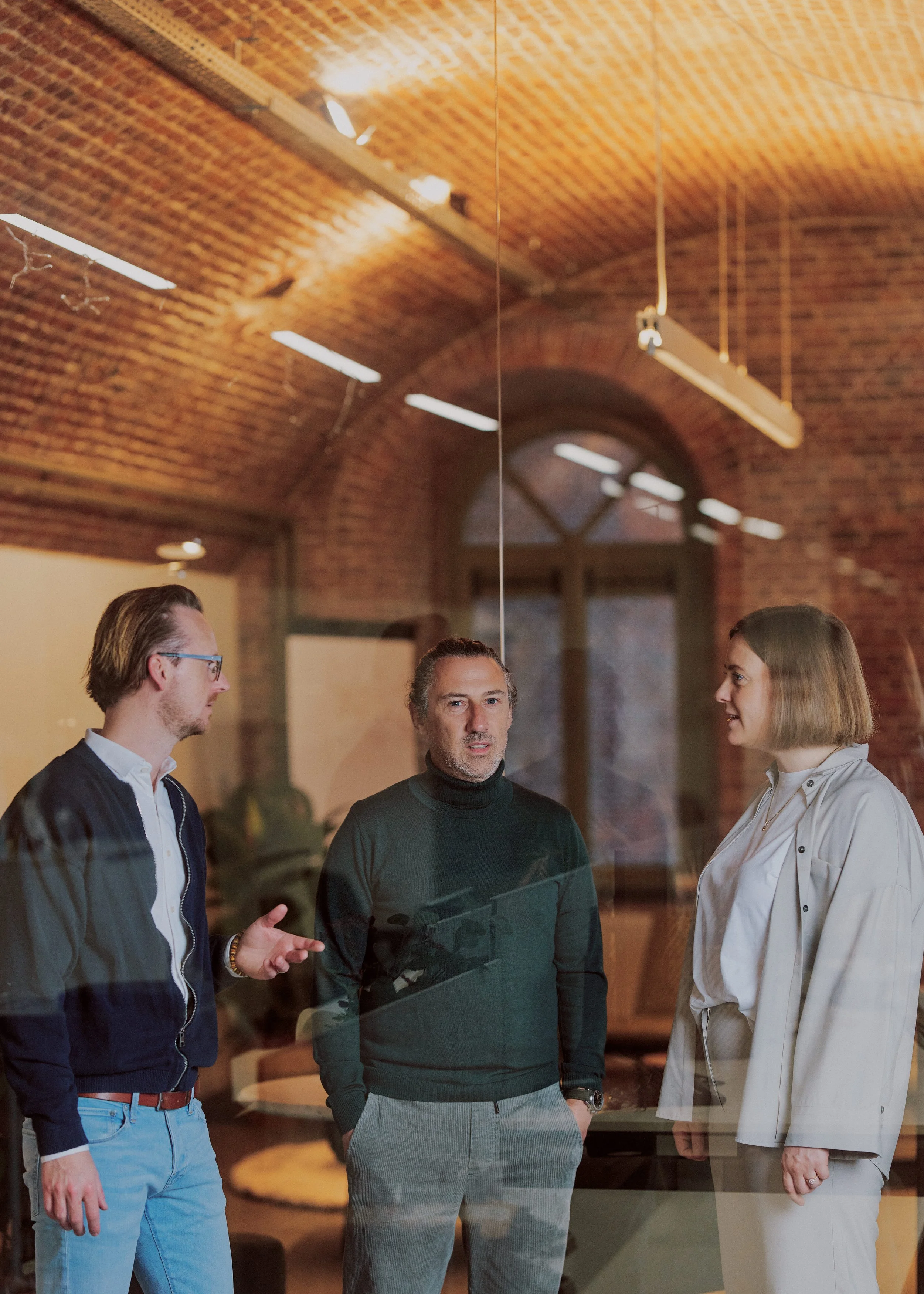 Four people standing and talking in a room with a brick ceiling and large arched window in the background.