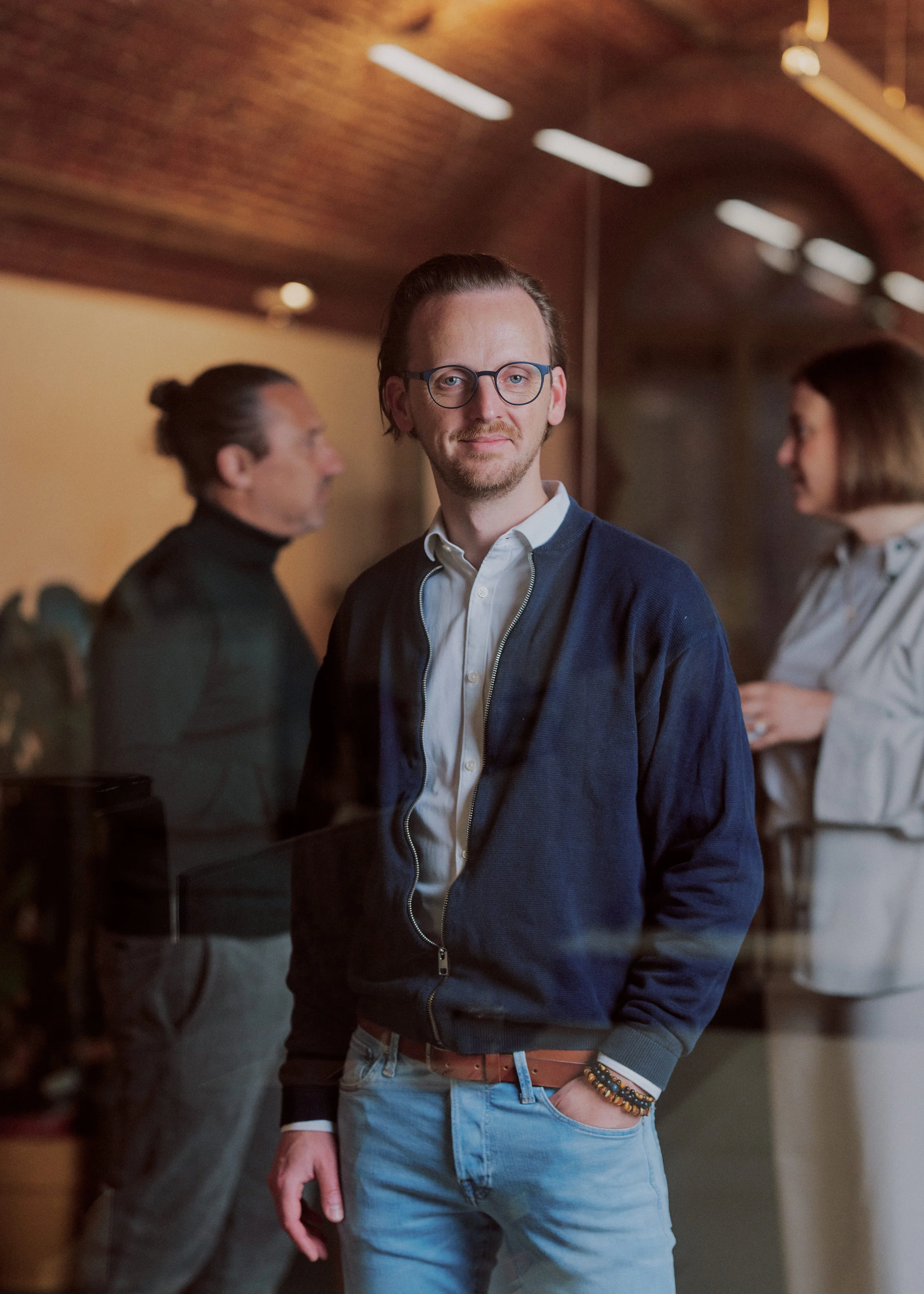 A young man with glasses and a beard smiling confidently in a modern indoor setting. He is wearing a white shirt, navy blue zip-up jacket, light blue jeans, and has bracelets on his wrist. Two blurred women are in the background, one with a bun hairstyle and the other with shoulder-length hair, engaging in conversation.
