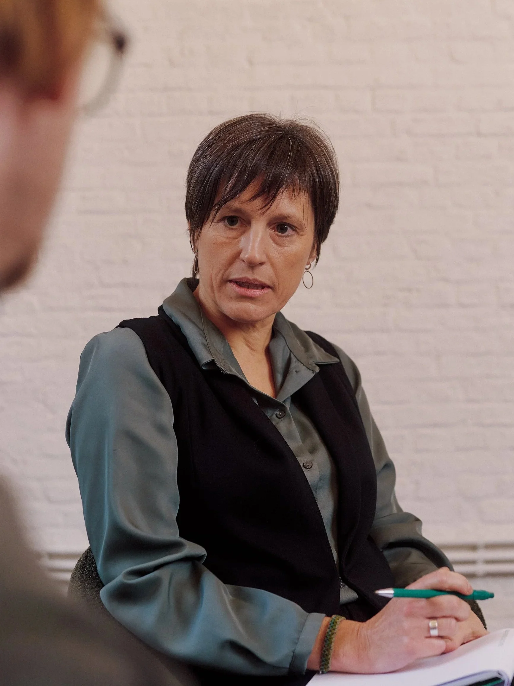 A woman with short dark hair and earrings, wearing a gray shirt and black vest, sitting at a table with a pen in her hand, in front of a white brick wall.