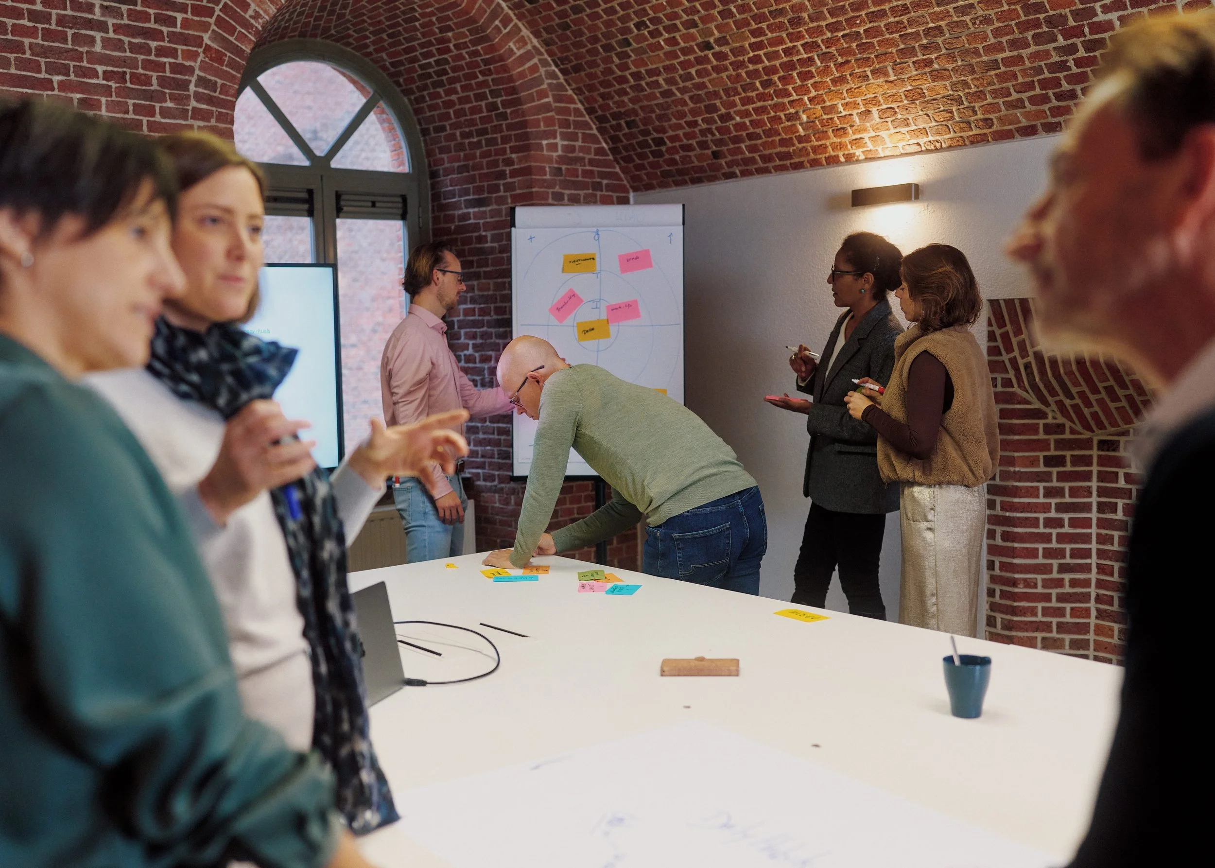 Group of people in a meeting room with a brick ceiling, some standing and some leaning over a white table, discussing and writing on a whiteboard with sticky notes. A large window is in the background.