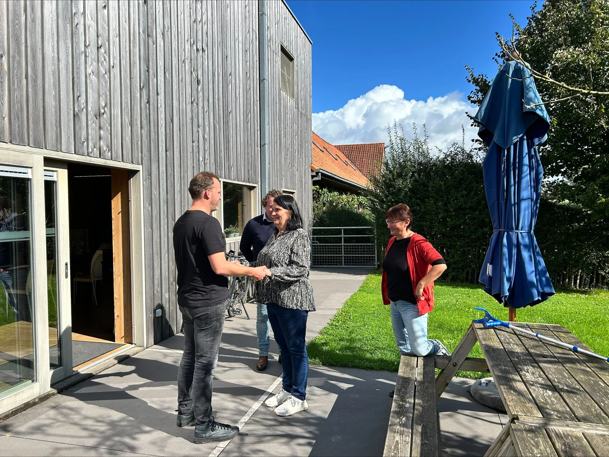 Four people standing outside by a modern building with a grey wooden exterior, blue sky with clouds, a blue umbrella, and a wooden picnic table.