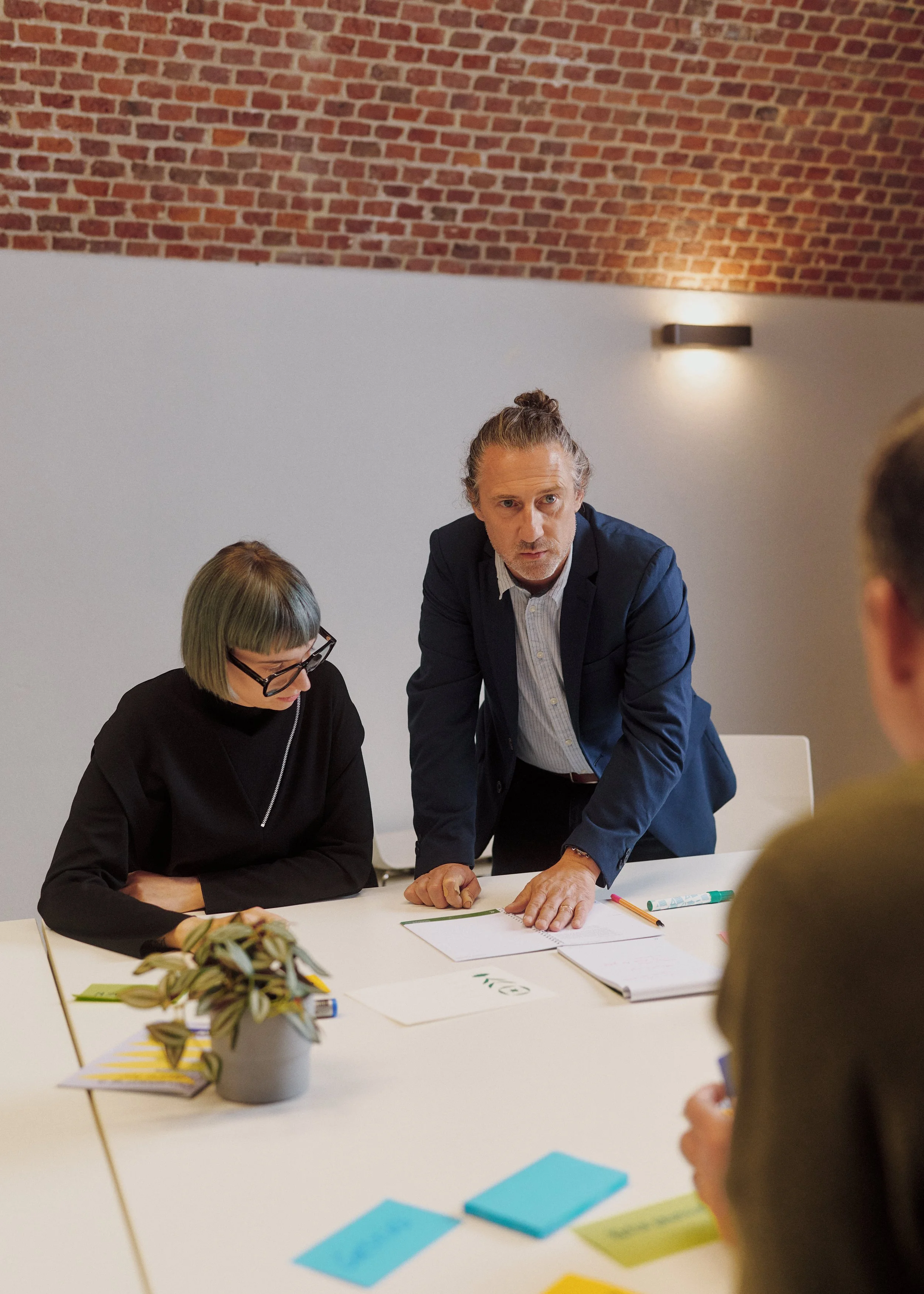 Four people are in a meeting room with a white wall and a brick ceiling. A man with gray hair, wearing a dark blue blazer and white shirt, is standing and speaking while a woman with short blonde hair, glasses, and a black jacket is sitting and listening. Two more people are partially visible, one with blonde hair and another with brown hair, sitting at the table. The table has notebooks, colorful pens, sticky notes, and a potted plant.