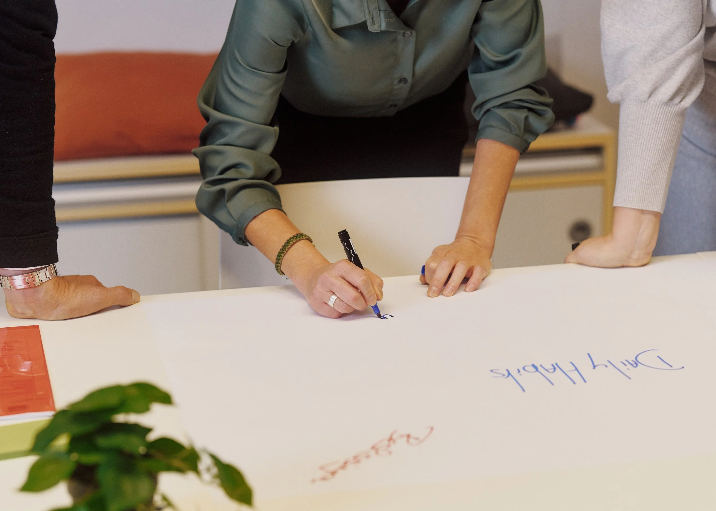 People are gathered around a table, signing or writing on a large sheet of paper. One person is holding a blue marker, and there are colorful sticky notes in the foreground. The setting appears to be a collaborative or meeting space.