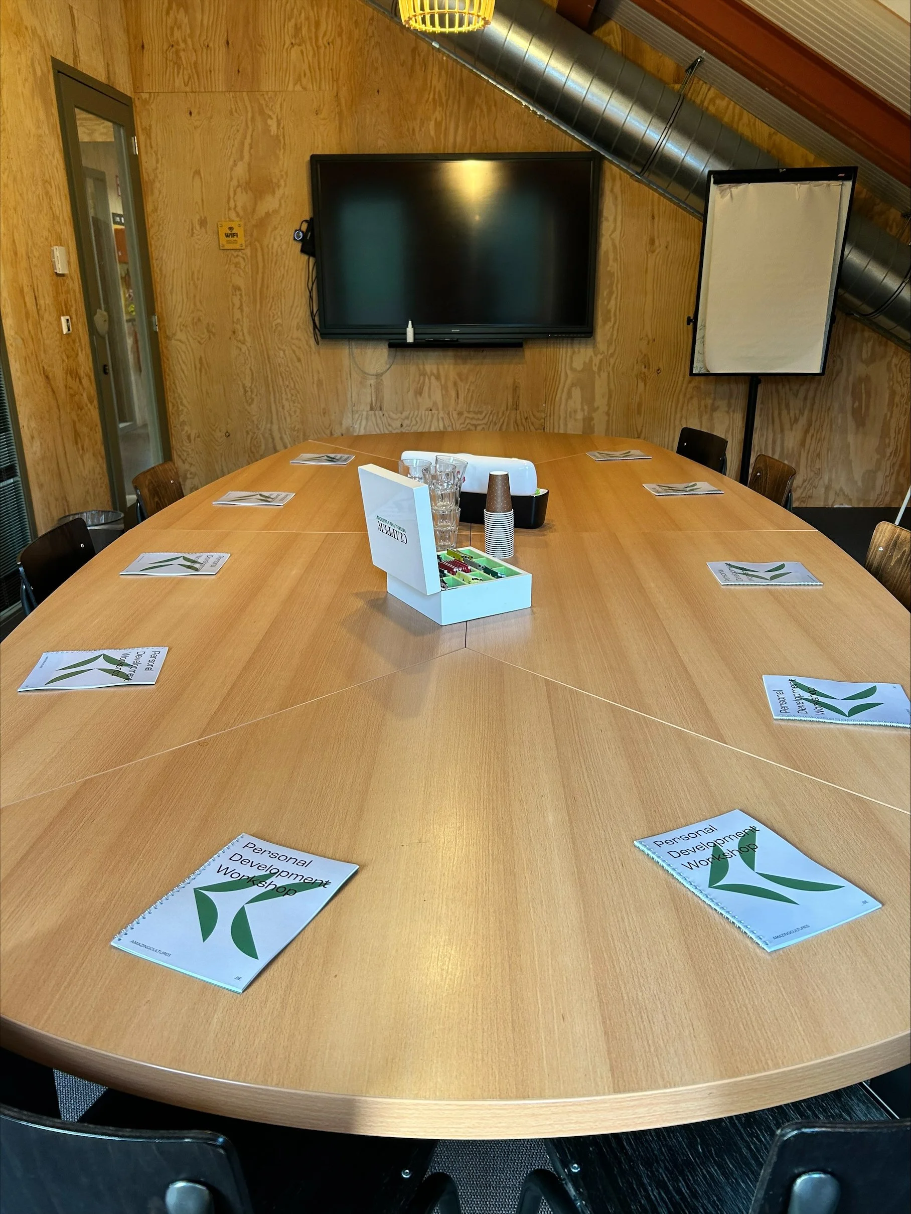 A round conference table with several booklets titled 'Personal Development Workshop,' some glasses, paper cups, a box of snacks, and a sanitizer bottle on top. The background features a wall with a flat-screen monitor, a window, and a softbox light, indicating a workshop or meeting room.